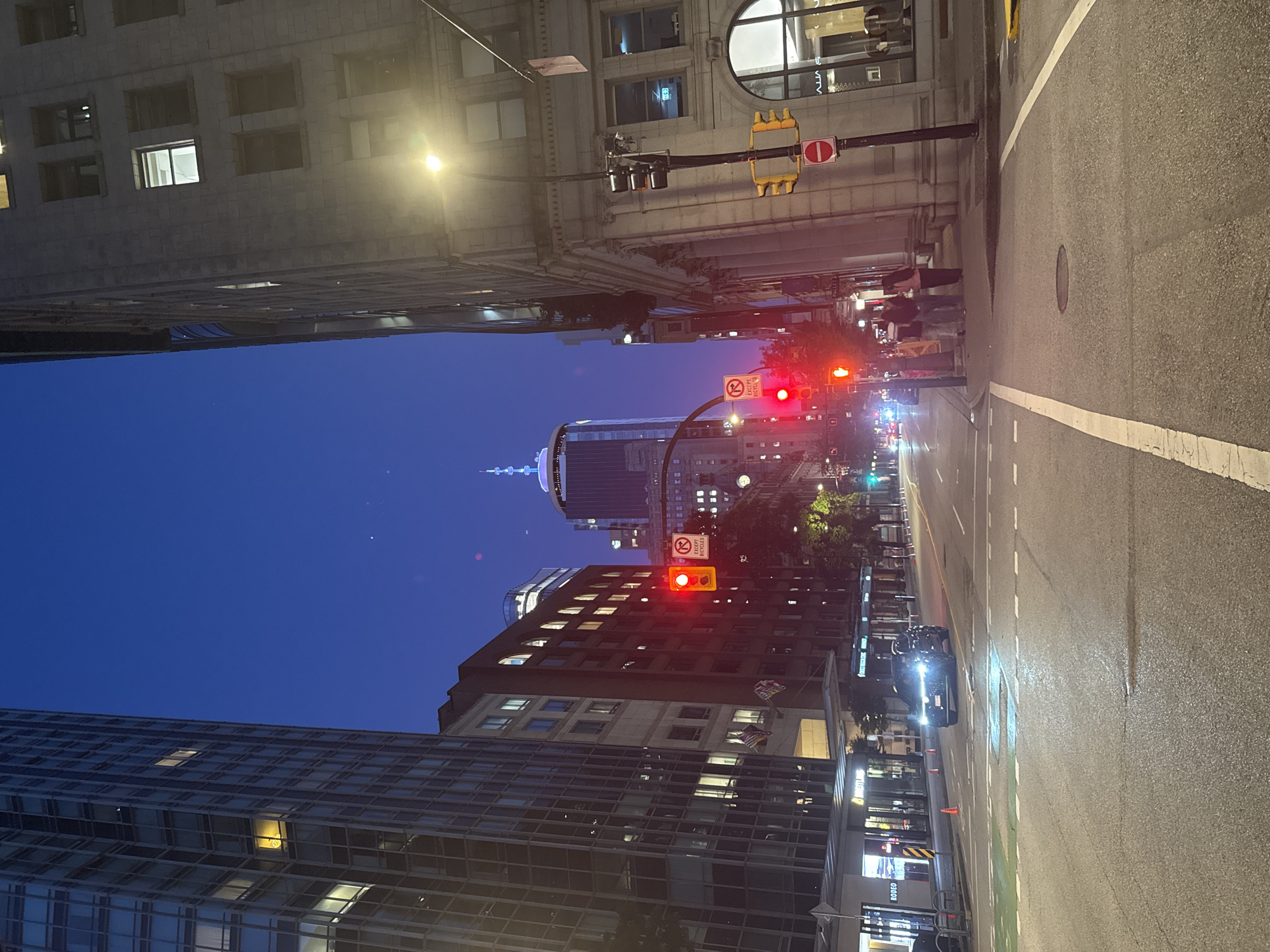 The image captures a street scene in downtown Vancouver, Canada, at dusk. The sky is a deep twilight blue, transitioning from the daylight to the night. Tall buildings flank the street on both sides. On the left, a modern glass-covered skyscraper dominates the view, its windows illuminated with warm interior lights. Adjacent to it is a building with a more traditional facade, also showing interior lighting.
On the right, an older, stone-faced building stands, its architectural details visible under the streetlights. A traffic light at the intersection is red, and a "No Parking Except Bicycles" sign is visible. Further down the street, another traffic light shows red, and another "No Parking Except Bicycles" sign is present.
In the distance, a prominent skyscraper with a distinct illuminated spire stands out against the sky. Several cars are on the road, with headlights illuminating the pavement. A dark-colored SUV is in the foreground, stopped at the red light. A group of four people are visible on the sidewalk near the right side of the street, appearing to be pedestrians waiting to cross.
The street itself is asphalt, with white lane markings and crosswalks. The atmosphere is urban and busy, with the city lights starting to twinkle as night approaches. The overall impression is one of a typical evening in a bustling downtown core.