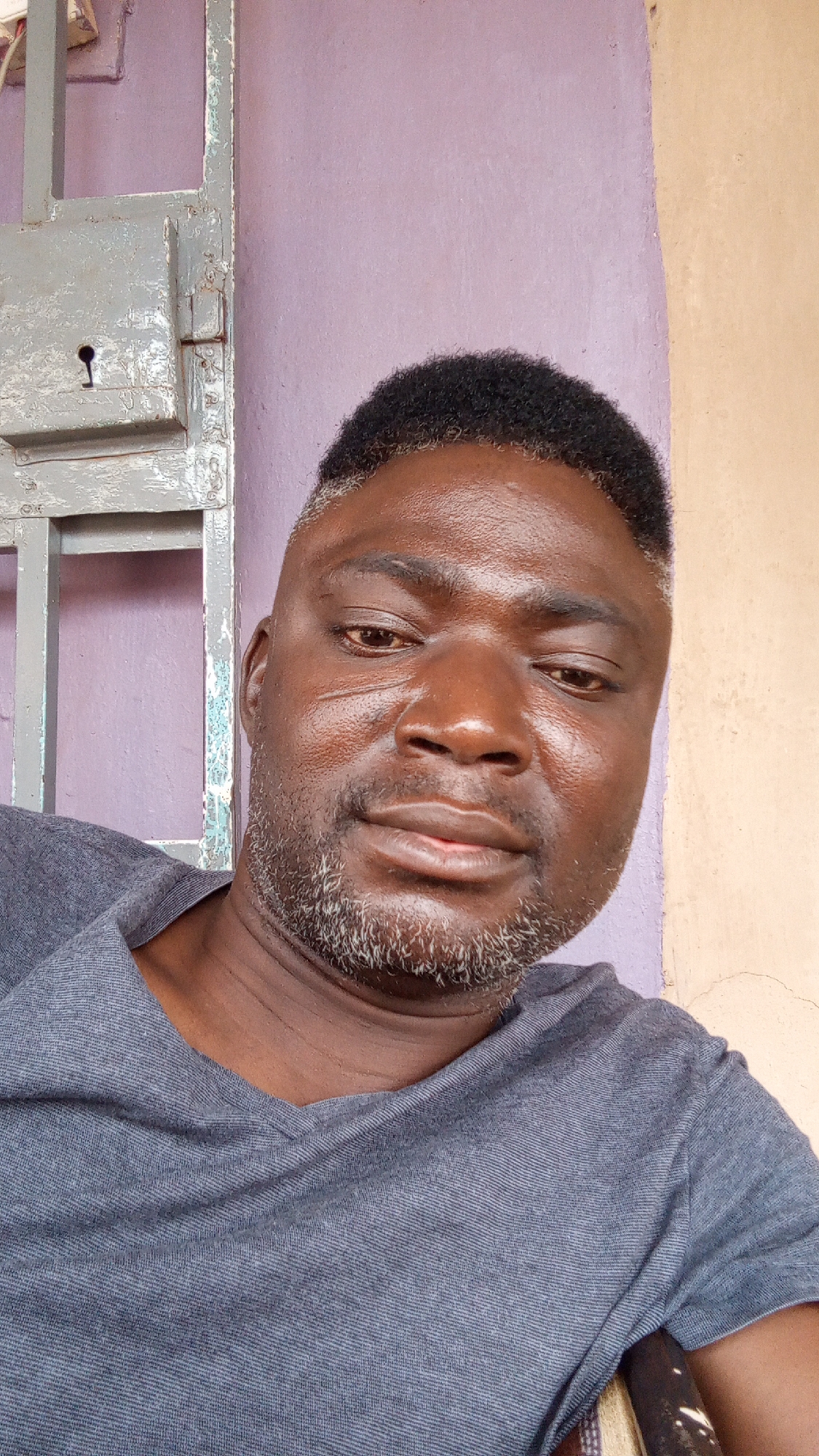 An adult Black man with short, dark hair and a salt-and-pepper beard is shown in a close-up head and shoulders shot. He wears a gray T-shirt. The background consists of a purple wall with a metal gate with a keyhole to the left and a tan wall to the right. The photograph appears to have been taken indoors. Sabon Tasha, Nigeria, is not identifiable in the image.