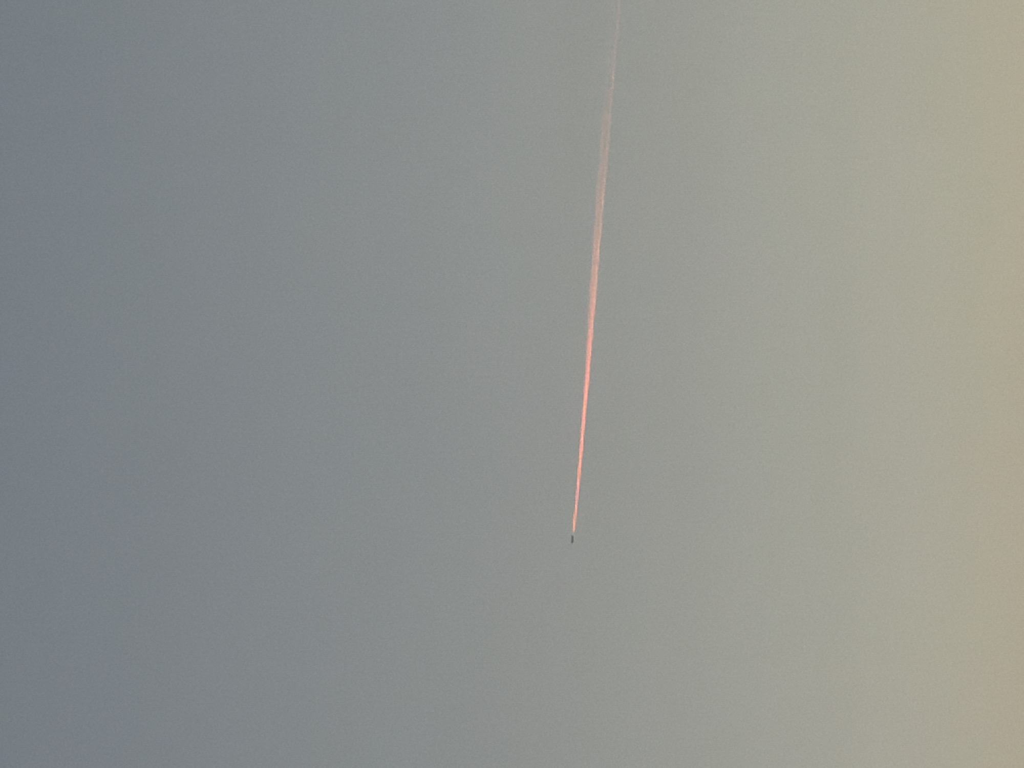 A distant airplane is visible in the sky, leaving a reddish contrail behind it. The airplane is a small, dark silhouette against the pale sky. The contrail is a thin, bright line that stretches horizontally across the image. The sky is a gradient of gray and pale yellow, indicating it may be early morning or late evening. The image appears to be taken in Leudelange, Luxembourg, as per the user's request.