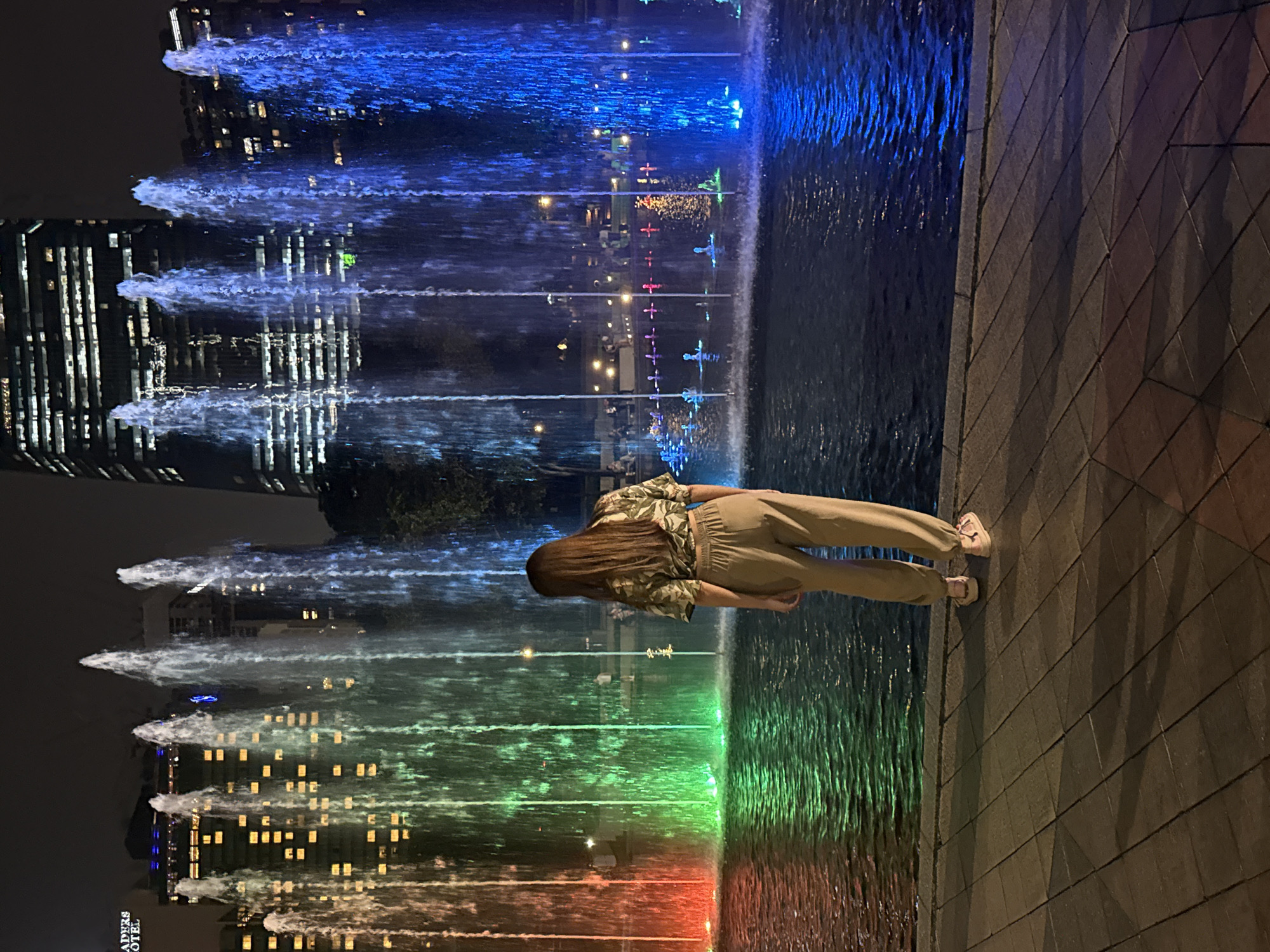 The image displays a vibrant nighttime scene centered around a colorful water fountain display. In the foreground, a woman stands with her back partially to the viewer, observing the spectacle. She has long, straight dark hair and is wearing a patterned short-sleeved shirt, light-colored wide-leg pants, and light-colored sneakers. She is standing on a paved surface, likely a promenade or plaza.
The main subject is a large water fountain system where multiple jets of water shoot upwards, illuminated by various colored lights. The colors transition across the fountain, featuring bright blues, greens, and reds, creating a dynamic and visually appealing show. The water's surface also reflects the colored lights and the surrounding environment.
In the background, several tall, modern buildings are visible against the dark night sky. Their numerous windows are lit, creating a grid of light. These building lights are also reflected in the water of the fountain, adding depth to the scene. The text "TRADERS HOTEL" is visible on one of the buildings in the lower left portion of the image, providing a location cue for an urban setting like Kuala Lumpur. The time of day is clearly night, indicated by the dark sky and reliance on artificial lighting for illumination. The activity is that of observing a public fountain display.