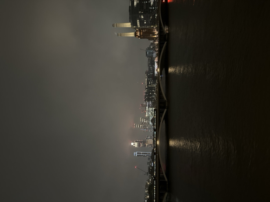 A nighttime cityscape stretches along a river's edge, illuminated by scattered lights. The river reflects these lights, creating streaks of brightness. A bridge spans the river, connecting the two sides of the city. Prominent buildings, including Battersea Power Station, are visible in the distance, their silhouettes contrasting against the dark sky. The overall mood is somber and atmospheric, with a sense of urban quietude.