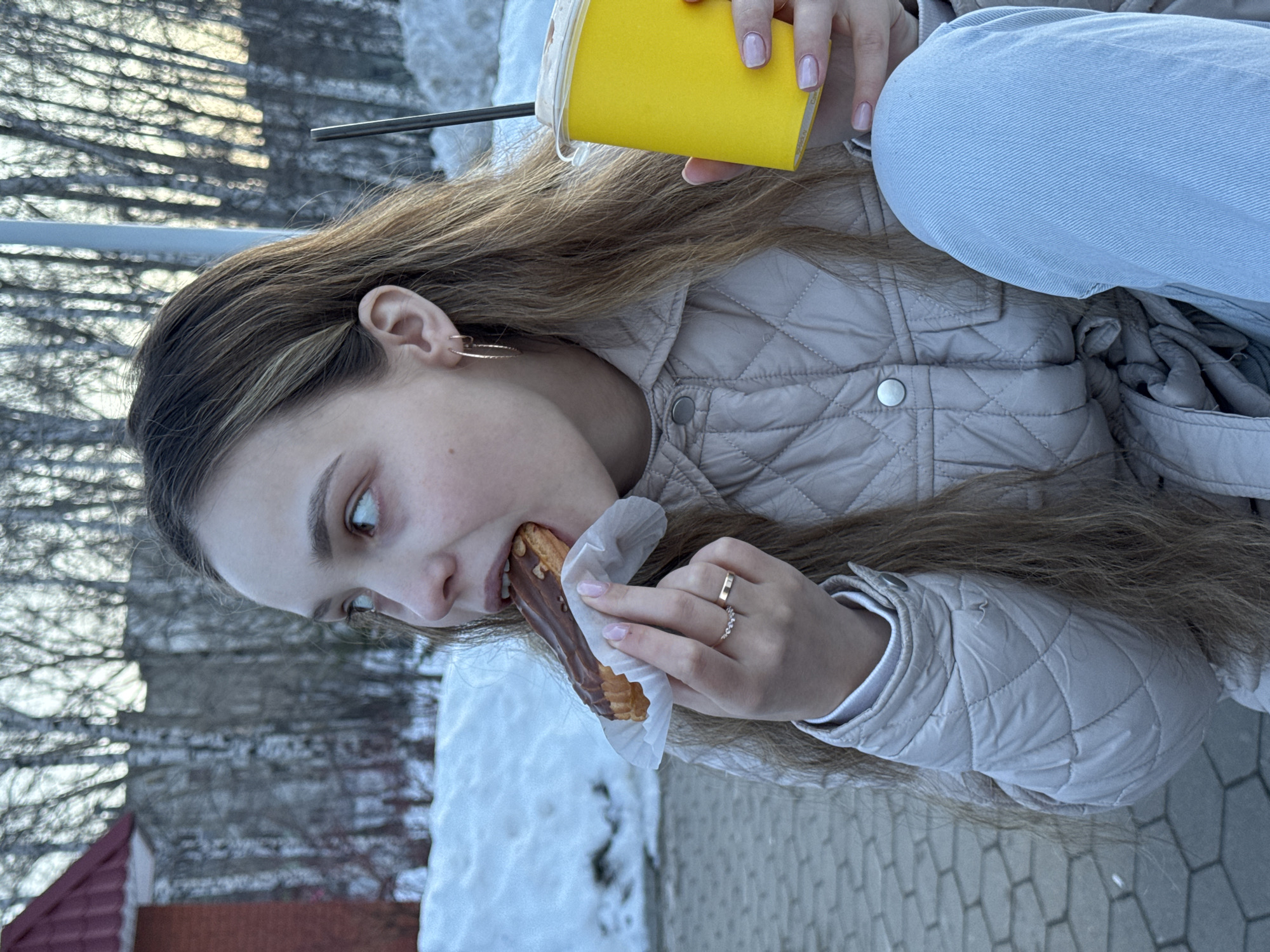 A young female is captured outdoors in Chaikovsky, Russia, in the act of consuming food and drink. She possesses long, light brown hair with lighter highlights and is observed looking upwards and to her right. Her left hand holds a chocolate-glazed eclair, partially bitten, wrapped in white paper. Simultaneously, her right hand grips a bright yellow disposable cup with a white lid and a dark straw. She is attired in a light beige quilted jacket and light blue jeans. Visible accessories include gold-colored hoop earrings in her right ear and two rings on her left ring finger, one thin gold-colored and one with small stones. Her fingernails exhibit light pink polish. The ground where she is positioned is paved with hexagonal gray blocks. The background features patches of snow, a partial view of a red-roofed structure to the left, and numerous bare deciduous trees, indicating a cold weather season. The scene is illuminated by natural light.