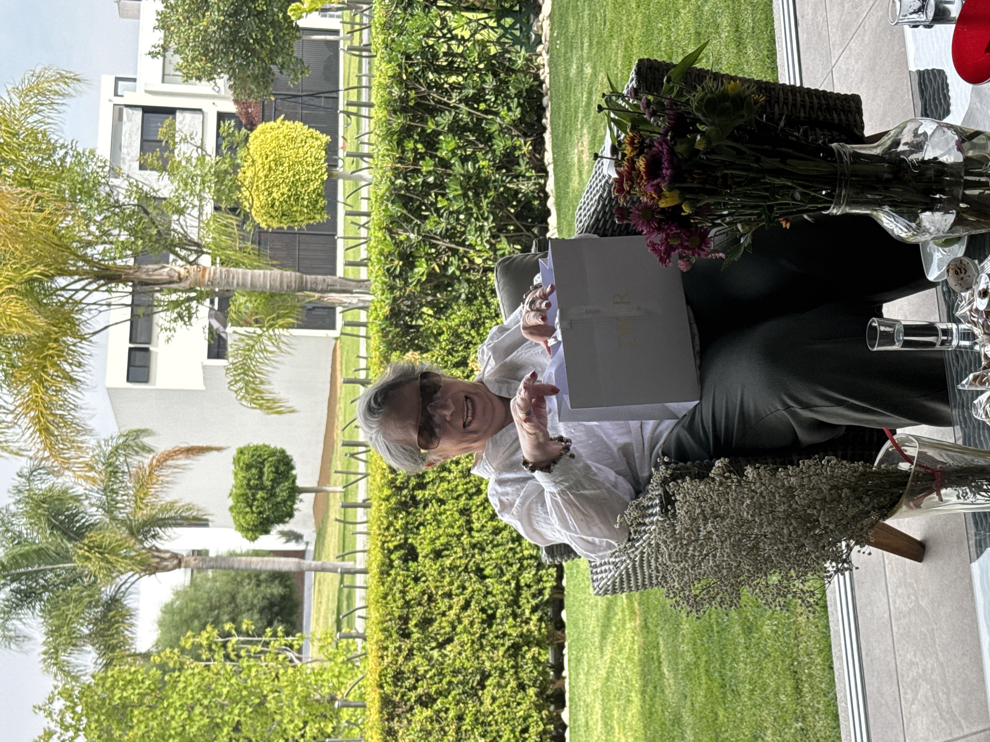 The image shows an older woman sitting outside in what appears to be a backyard or patio. She is holding a white shopping bag with the word "DIOR" printed on it. She is wearing sunglasses and a white blouse. There are flowers on the table beside her, both in a vase and arranged in a bouquet. The background features a well-maintained green lawn, hedges, trees, and a building in the distance. The location is San Bernardino Tlaxcalancingo, Mexico, as indicated by the image details.