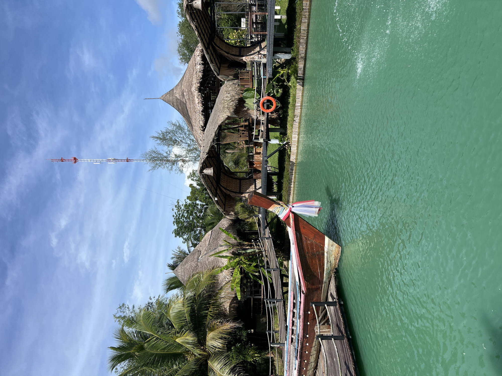 The image depicts a scenic view of a waterfront setting with traditional Thai architecture and a longtail boat. The scene is dominated by a large body of calm, turquoise water, likely a river or bay, with ripples suggesting a gentle current or a fountain in the background. Adjacent to the water is a wooden dock leading to a longtail boat, its prow adorned with colorful fabric streamers.
Behind the dock, several huts with conical, thatched roofs are visible, interspersed with lush green tropical foliage, including palm trees. The structures appear to be part of a resort or restaurant, designed in an open-air style. A life preserver ring hangs from a post near one of the huts. In the distance, a tall, red and white telecommunications tower pierces the bright blue sky, which is dotted with wispy white clouds.
The overall impression is one of a sunny day in a tropical location. The light suggests it's daytime, likely mid-morning or afternoon. The vibrant colors of the water, greenery, and boat, along with the clear sky, evoke a peaceful and inviting atmosphere. There are no people visible in the image, focusing the viewer's attention on the natural beauty and architectural elements of the place. The location cues, such as the tropical vegetation and the longtail boat, combined with the provided location context of Tambon Pa Kae Bo Hin, Thailand, strongly suggest the setting is in Thailand. There is no visible text in the image.