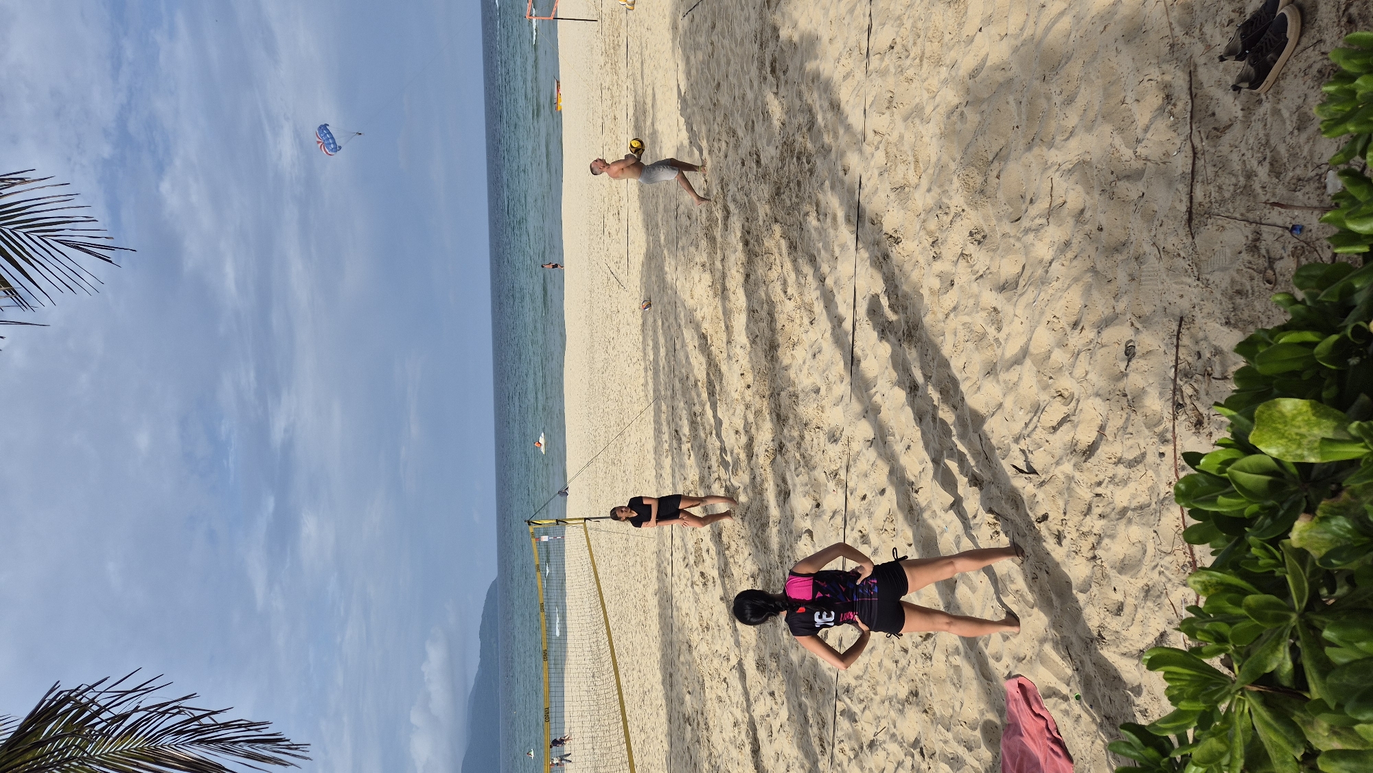 The image depicts a beach volleyball game on a sandy beach in Đà Nẵng, Vietnam. Three individuals are visible on the court. On the left, a person wearing a black and pink athletic top and black shorts stands with their hands on their hips, facing away from the camera. In the middle, a person in a black t-shirt and black shorts stands with their arms crossed. On the right, a man wearing grey shorts is in motion, holding a yellow volleyball. A volleyball net spans the width of the court. In the background, the ocean meets the horizon under a cloudy sky. Several other people are scattered on the beach and in the water, and a parasailer is visible in the sky. Lush green foliage frames the bottom of the image. The scene appears to be a recreational activity on a typical beach day in Đà Nẵng.