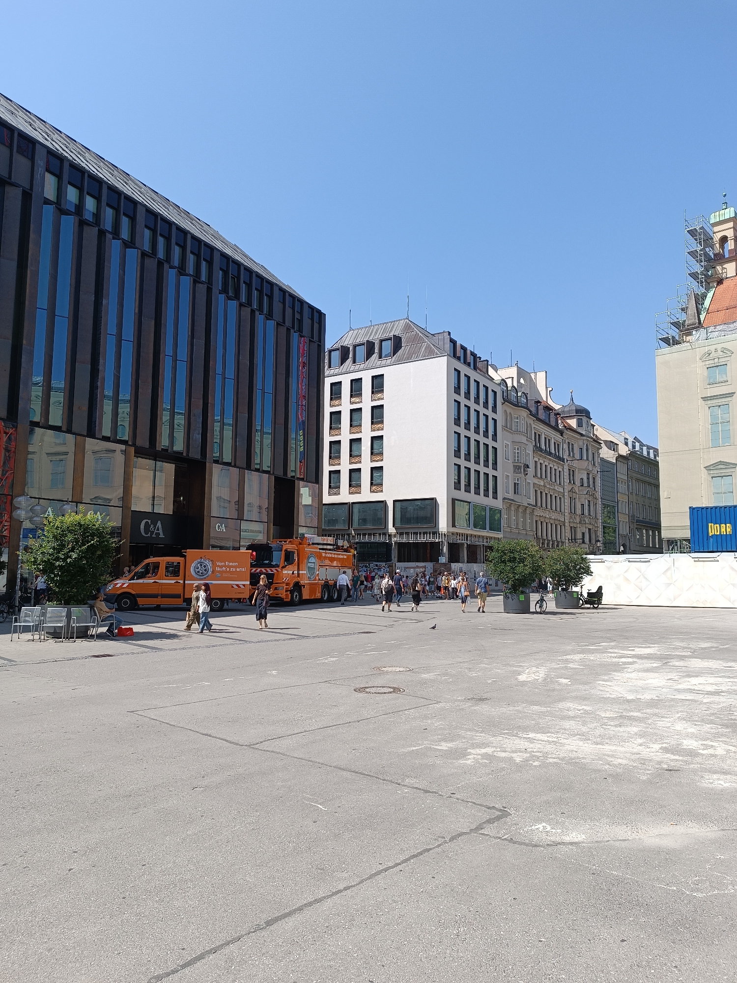 This image depicts a street scene in Munich, Germany, specifically in the area of Marienplatz. The foreground shows a wide, paved pedestrian area with some manhole covers and a few scattered white chairs.
To the left, a modern building with a dark, vertical facade of glass and metal dominates the scene. The "C&A" logo is visible on the lower part of this building, indicating a retail store. In front of the building, a green tree provides a splash of color.
Adjacent to the C&A building, an orange van and a larger orange vehicle resembling a fire truck are parked. The van has lettering on its side, including "Von Ihnen hofft zu uns!" and a circular logo. People are walking on the sidewalk in front of these vehicles and the building.
Further into the background, a series of older, multi-story buildings with traditional European architecture line the street. These buildings have varied facades and rooflines, some featuring dormer windows and decorative elements. One building has scaffolding around its upper section, suggesting ongoing construction or renovation. To the far right, a blue shipping container with "DORA" written on it is visible, along with more construction elements. The sky is clear and blue, indicating a sunny day.