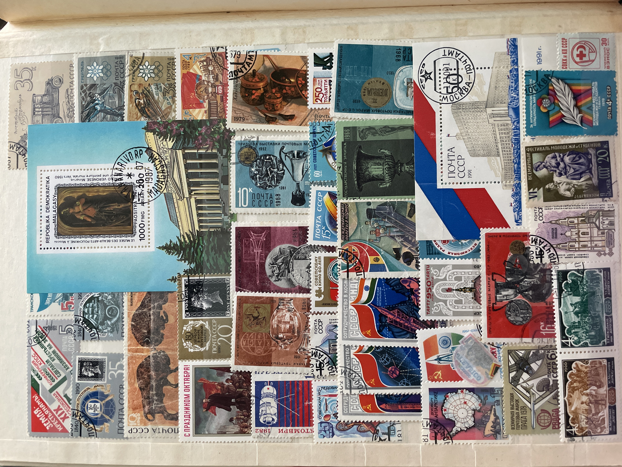 The image is a close-up overhead view of a page from a stamp album filled with postage stamps from the Soviet Union and other countries. The stamps depict various themes including historical events, art, architecture, and space exploration.
One stamp, located in the upper center of the image, features a depiction of the Moscow State University, a prominent landmark in the city of Moscow, Russia. The stamp, labeled "ПОЧТА СССР 1991", also shows a postmark with "МОСКВА - ПОЧТАМТ" indicating its origin. While the university is located in Moscow, there is no indication in the image that this location is related to the city of Сокол, Russia.
The page is organized with stamps arranged in rows and columns, some overlapping. The background of the album page appears to be a light-colored paper, possibly off-white or cream. The lighting suggests an indoor setting with diffuse light. No people are visible in the scene, and the focus is entirely on the collection of stamps. The stamps themselves vary in color, size, and design, showcasing a diverse range of imagery.