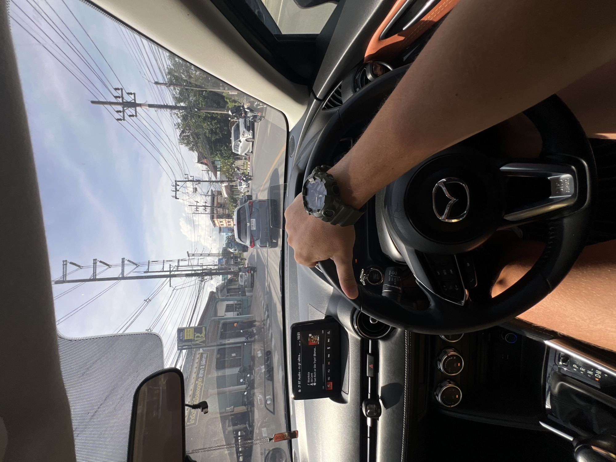 This media file is a first-person perspective shot from inside a car, likely a Mazda, as indicated by the logo on the steering wheel. The driver's arm, adorned with a green digital watch, is visible resting on the steering wheel. The scene outside the car shows a street lined with buildings and utility poles with numerous wires. Traffic appears to be present, with cars and scooters visible on the road. The sky is bright and appears to be daytime, with clear weather conditions. A car's infotainment system is displaying music information, showing "BT Audio - rs gt ultra...." and song details. Visible text includes "Al Ritz", "AR", and Thai script that translates to "Secondhand tire warehouse Phuket Branch 2 (Baan Krok)". The overall impression is of a journey through a town or city during the day.