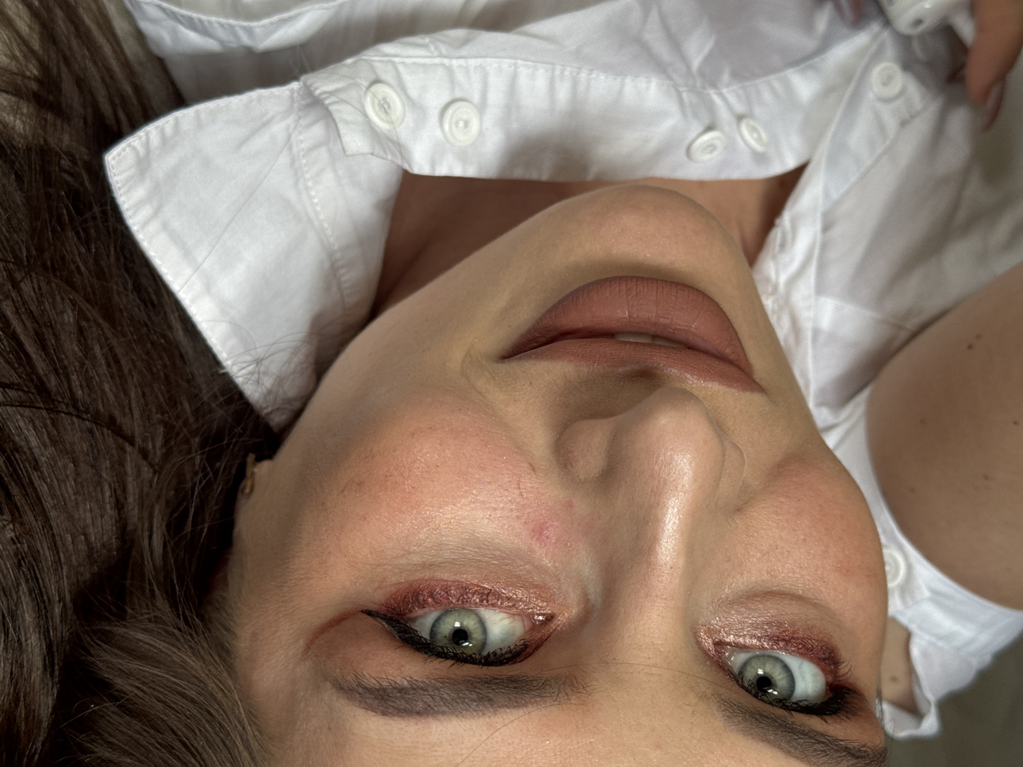 A close-up, low-angle portrait captures a woman with her head tilted back, looking directly at the camera with a gentle, pleasant smile. The image suggests she is lying down or reclined, with the camera positioned above her.
The subject is a woman with fair skin, prominent cheekbones, and dark brown hair, part of which is visible on the left side of the frame. Her eyes are a striking green-grey, enhanced with a shimmery reddish-pink eyeshadow, precise black winged eyeliner, and dark mascara. Her eyebrows are well-defined and filled in. Her lips are full, colored with a matte nude or light brown lipstick, and a slight hint of a smile reveals the top row of her teeth. There is some minor skin redness or blemishes visible on her forehead and cheek.
She is wearing a crisp white button-up shirt, partially unbuttoned, revealing a glimpse of her neck and chest area. The fabric shows subtle wrinkles and stitching details. Several white buttons are visible along the placket of the shirt, along with the structured collar.
The overall impression is a soft, intimate portrait. The time of day and weather are not discernible from the image. The location context provided is Rusalevka, Russia. There is no visible text in the media file.