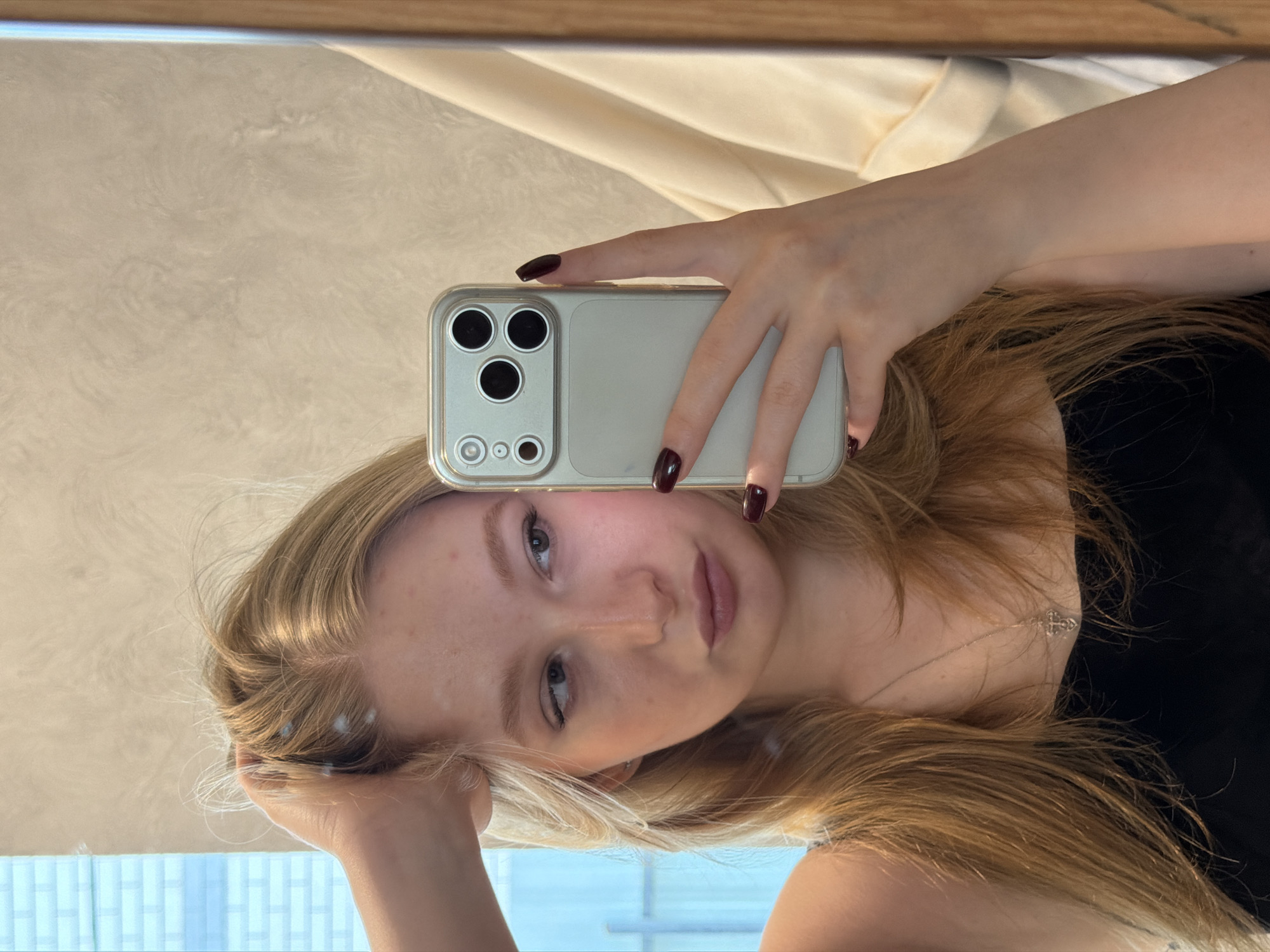 A fair-skinned young woman with blonde hair is taking a selfie in Rossiyskiy, Russia. She is holding a white smartphone with a multi-lens camera system. Her nails are painted a dark maroon color. A small silver necklace is visible around her neck. The background includes a beige wall and a window with blue and white curtains.