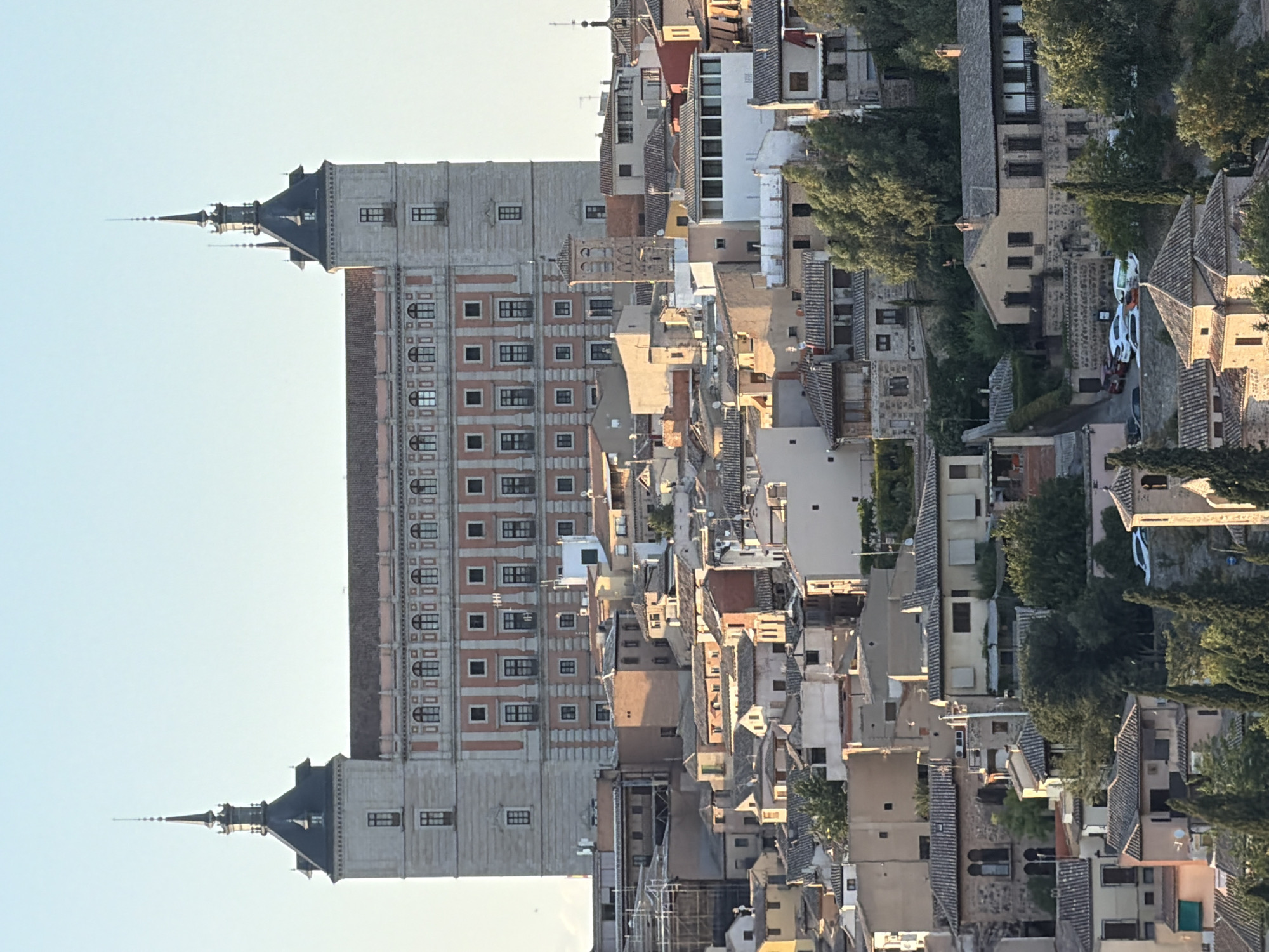 The image shows a panoramic view of the city of Toledo, Spain. In the background, the Alcázar of Toledo dominates the skyline, a large fortress palace with distinctive towers and a red-tiled roof. The building is flanked by smaller, tightly packed structures, typical of historic European cities, with a mix of architectural styles evident. The foreground and midground reveal a dense urban landscape of houses and buildings, many with red-tiled roofs, clustered on a hillside. The buildings are of varying sizes and colors, with some showing signs of age and wear. Trees and vegetation are interspersed throughout the cityscape, adding a touch of green to the otherwise muted tones of the buildings. The overall impression is of a historic and densely populated city with a prominent landmark at its center. The lighting suggests it is either early morning or late afternoon, with warm sunlight illuminating the buildings. There are no visible people in the image.