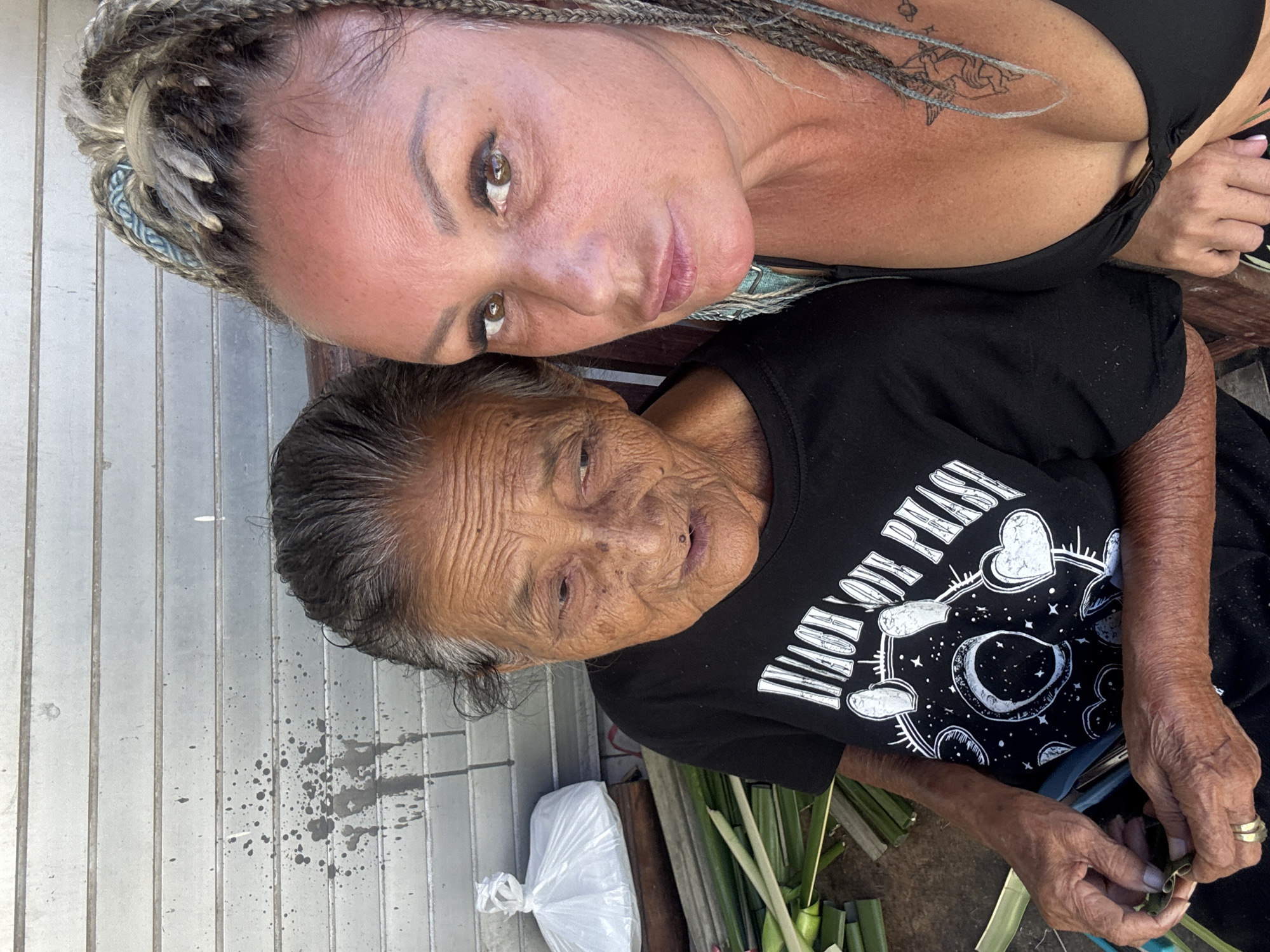 This image, set in Kuta, Indonesia, depicts two women in a close-up, rotated view, suggesting they are side-by-side. The lighting indicates a bright, sunny day outdoors.
The younger woman, positioned at the top, has light-colored skin, dark eye makeup, and full lips. Her hair is styled in blonde and grey braids, some secured with a light blue and white patterned fabric band. She wears a black top, possibly a bikini or tank top, which reveals a tattoo of a stylized creature (possibly a unicorn or pegasus) with stars on her upper chest/shoulder, and another small tattoo further up. She looks directly towards the viewer with a calm expression.
Below her is an older woman with wrinkled, darker skin and greyish-black hair. She wears a black t-shirt with a white graphic design. The design includes the text "LIVE ON PLANET", where the 'A' in 'PLANET' is stylized as a celestial body (moon/planet) surrounded by stars, and two hearts appear above it. Her hands, showing signs of age, are engaged in preparing or weaving green palm-like leaves or fronds. She has two rings on one finger. Her gaze is directed downwards, focused on her task.
In the background, behind the younger woman, is a light-colored, vertically slatted surface. Behind the older woman, to her left, is a white plastic bag, and to her right are numerous bundles of the green leaves she is working with, some already folded or cut. Dark splatters are visible on the slatted background near the older woman's head. The overall scene captures an intimate moment of intergenerational interaction and a traditional craft activity.