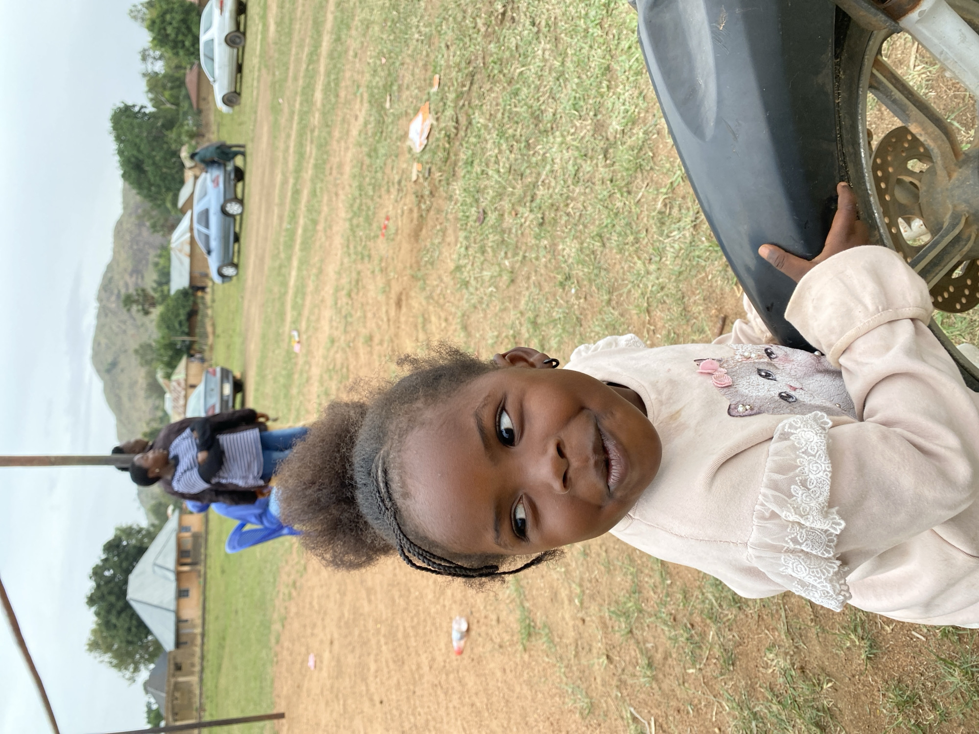 A young girl is close to the camera, smiling while holding onto the fender of a motorcycle. The setting appears to be outdoors, in a grassy field. Cars are parked in the background. People stand near a tent. The location is in Kuru Dungus, Nigeria.