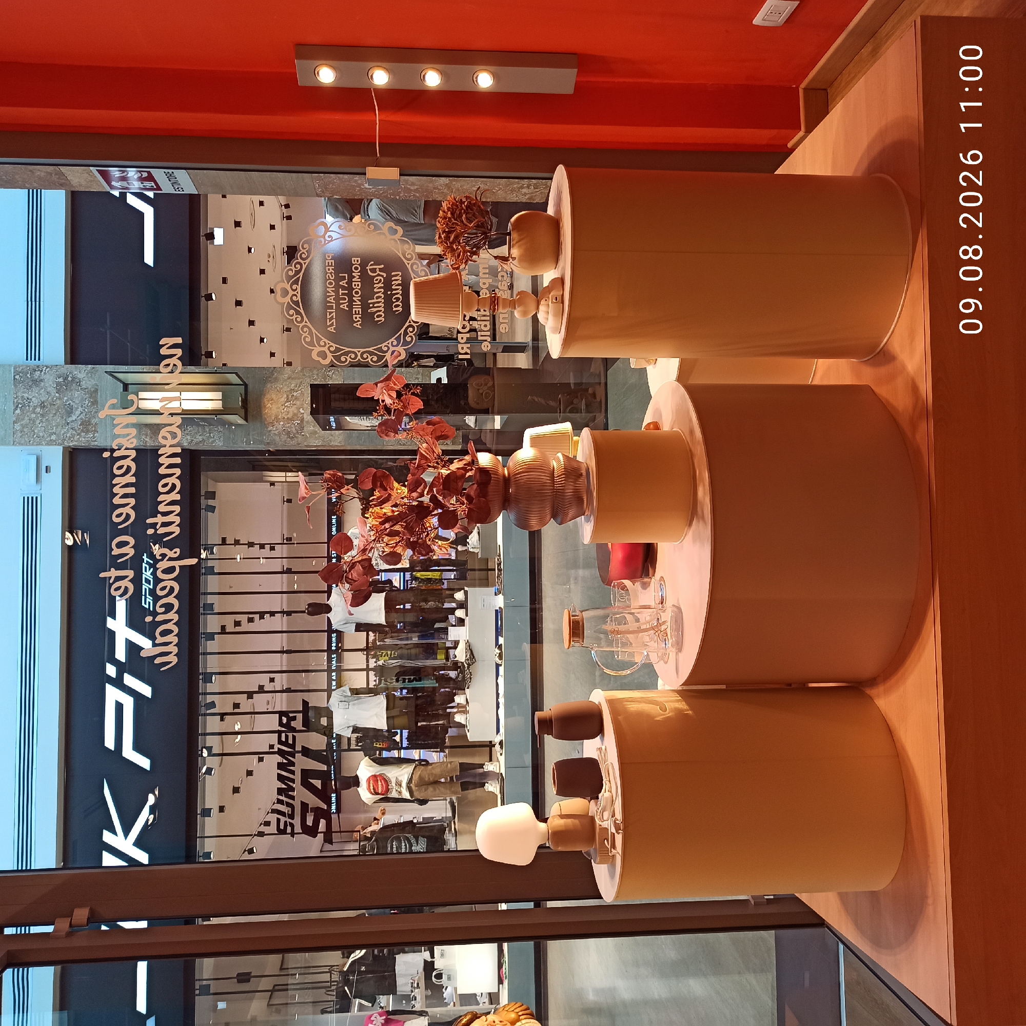 This is a photograph of a store window display in Molfetta, Italy, featuring a "SUMMER SALE" sign and mannequins dressed in casual attire. The display includes various decorative objects such as lamps, vases, and plants. In the foreground, there are cylindrical pedestals holding these items, with a glass pitcher and cups on one of them. In the background, through the glass, the interior of the store is visible with clothing racks and more mannequins. The overall lighting is artificial and somewhat dim, creating a warm ambiance. A date and time stamp "09.08.2026 11:00" is overlaid on the lower right portion of the image.