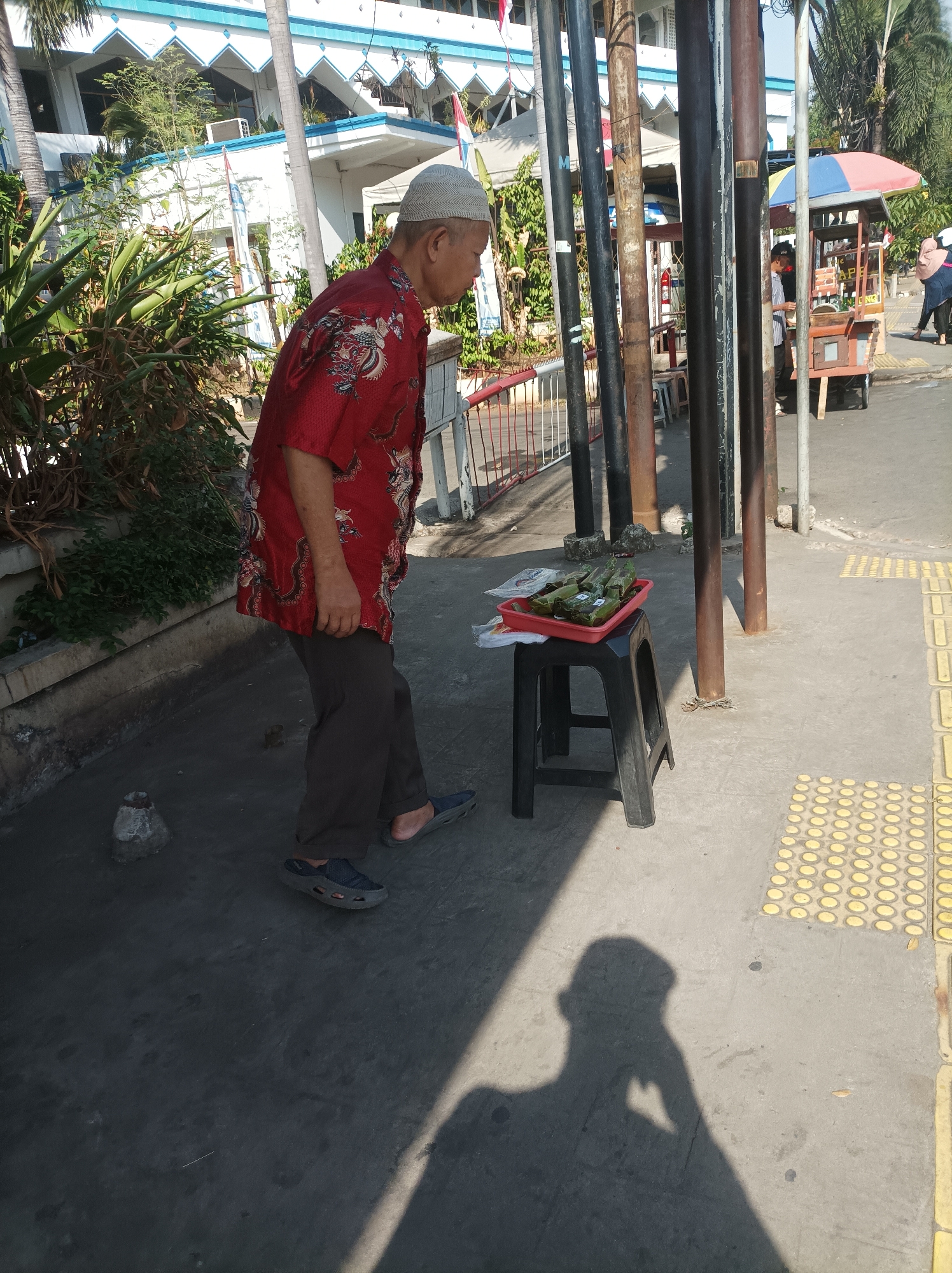 An elderly man wearing a red shirt with a dragon motif and a white skullcap is standing on a sidewalk. He is looking down at a red plastic tray filled with food items wrapped in banana leaves, which is placed on a black plastic stool. The man is wearing brown pants and dark sandals. The sidewalk is paved with concrete and has a textured yellow tactile paving system. In the background, a building with white and blue accents and trees can be seen. There are several utility poles on the sidewalk. To the right, a food cart with a colorful umbrella is visible, and two women are standing near it. The scene appears to be a street in South Jakarta, Indonesia, possibly near a mosque given the architectural style of the building. The lighting suggests it is daytime, with strong shadows cast by the man and the utility poles.