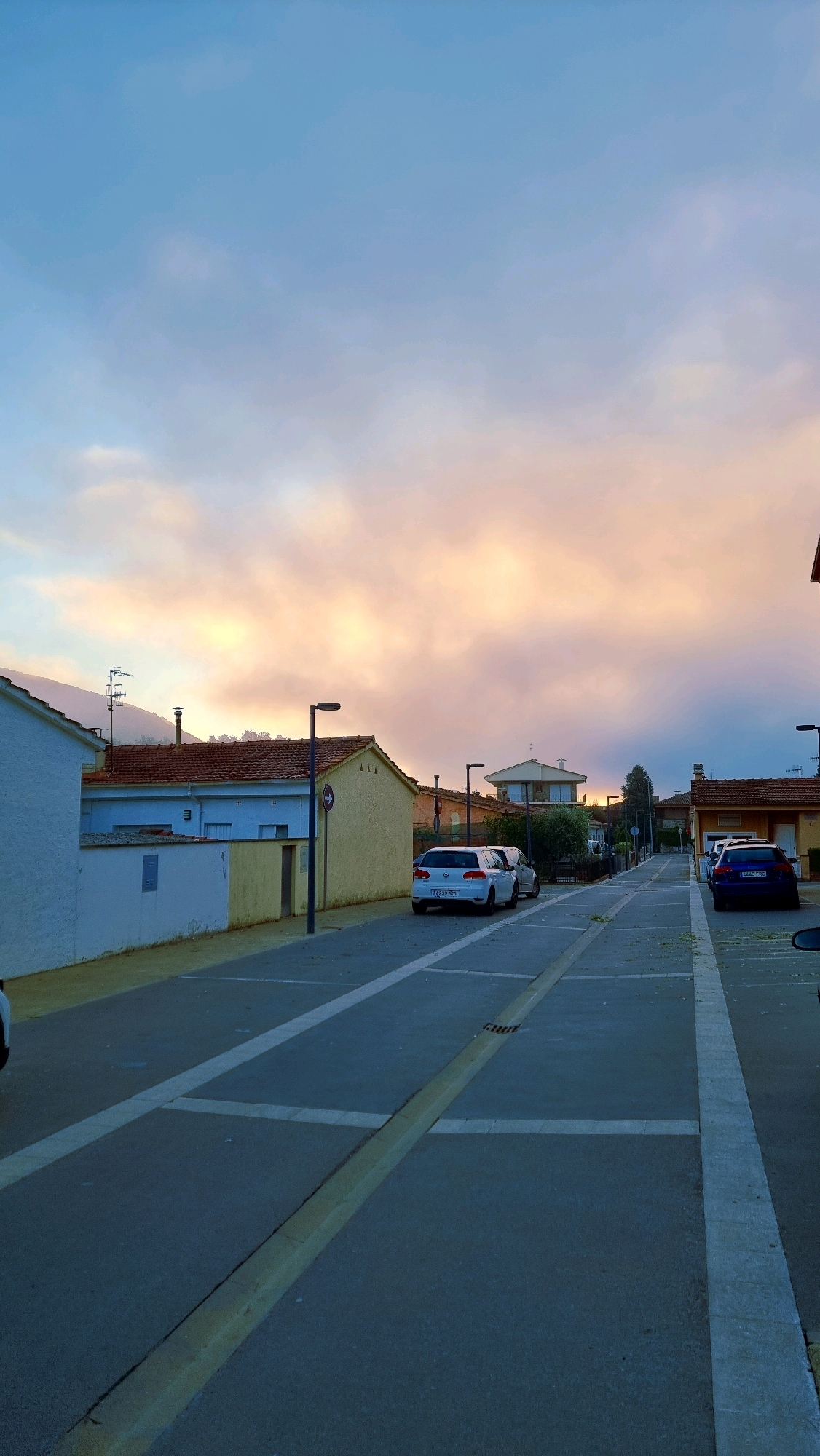 The image depicts a street scene in Sant Joan Les Fonts, Spain, under a twilight sky. The sky is a gradient of soft blues and pastel oranges, indicating either sunrise or sunset, with scattered clouds.
The street is paved and marked with white lines, suggesting a parking area or designated lanes. On either side of the street are residential buildings with terracotta roofs. The houses appear to be single-story or two-story structures with a mix of white, yellow, and light blue facades.
Several cars are parked along the street. On the left, a white hatchback is parked. Further down, another white hatchback is parked. On the right side of the street, a dark blue sedan is visible. Streetlights are positioned along the road.
In the background, more houses and some vegetation can be seen. The presence of antennas on one of the houses suggests a residential area. The overall atmosphere is quiet and serene, typical of a suburban or small-town environment during the transitional hours of the day.