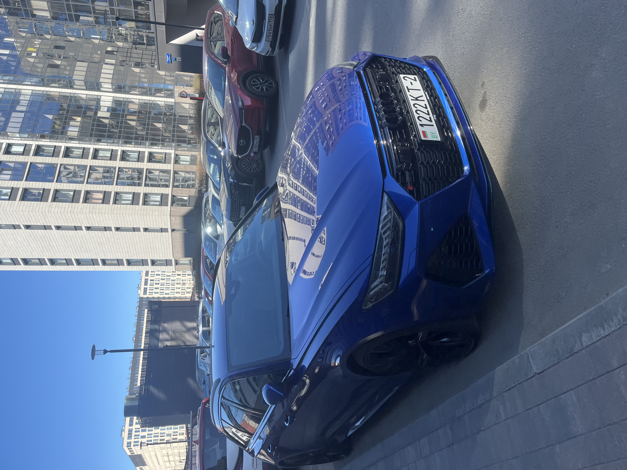 The image depicts a blue Audi RS3 parked on a city street. The car is facing the viewer at an angle, and its license plate, "1222 KT-2 BY," identifies its origin as Belarus. The surrounding environment is a modern urban setting, characterized by glass-fronted buildings, and several other cars parked nearby. The location is identified as Saint Petersburg, Russia, based on the reflections on the blue car’s hood. There are no visible people in the scene.