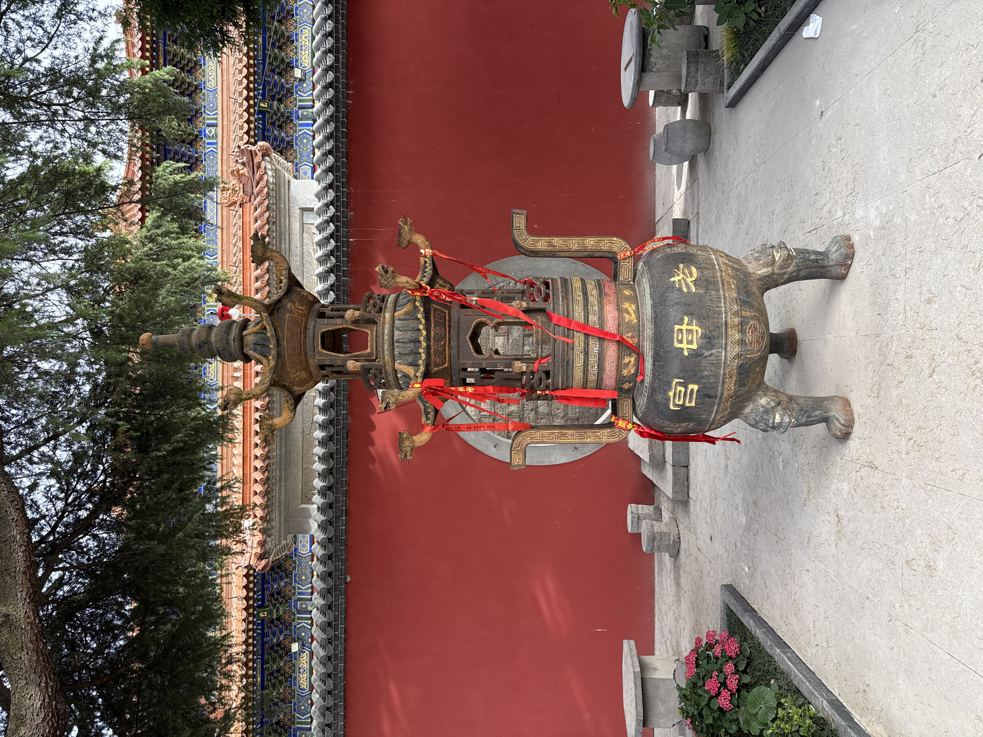 The image shows a large, elaborate bronze incense burner in an outdoor setting. The burner is decorated with intricate designs, including small dragon figures and bands of ornate patterns. Red ribbons are tied around the burner. Chinese characters are visible on the side. The burner sits on a stone-tiled area near a red wall with traditional Chinese architectural details. A tree is visible on the left side of the image and a flower bed with red flowers is visible on the bottom right. The picture was taken in Lintong District, China.
