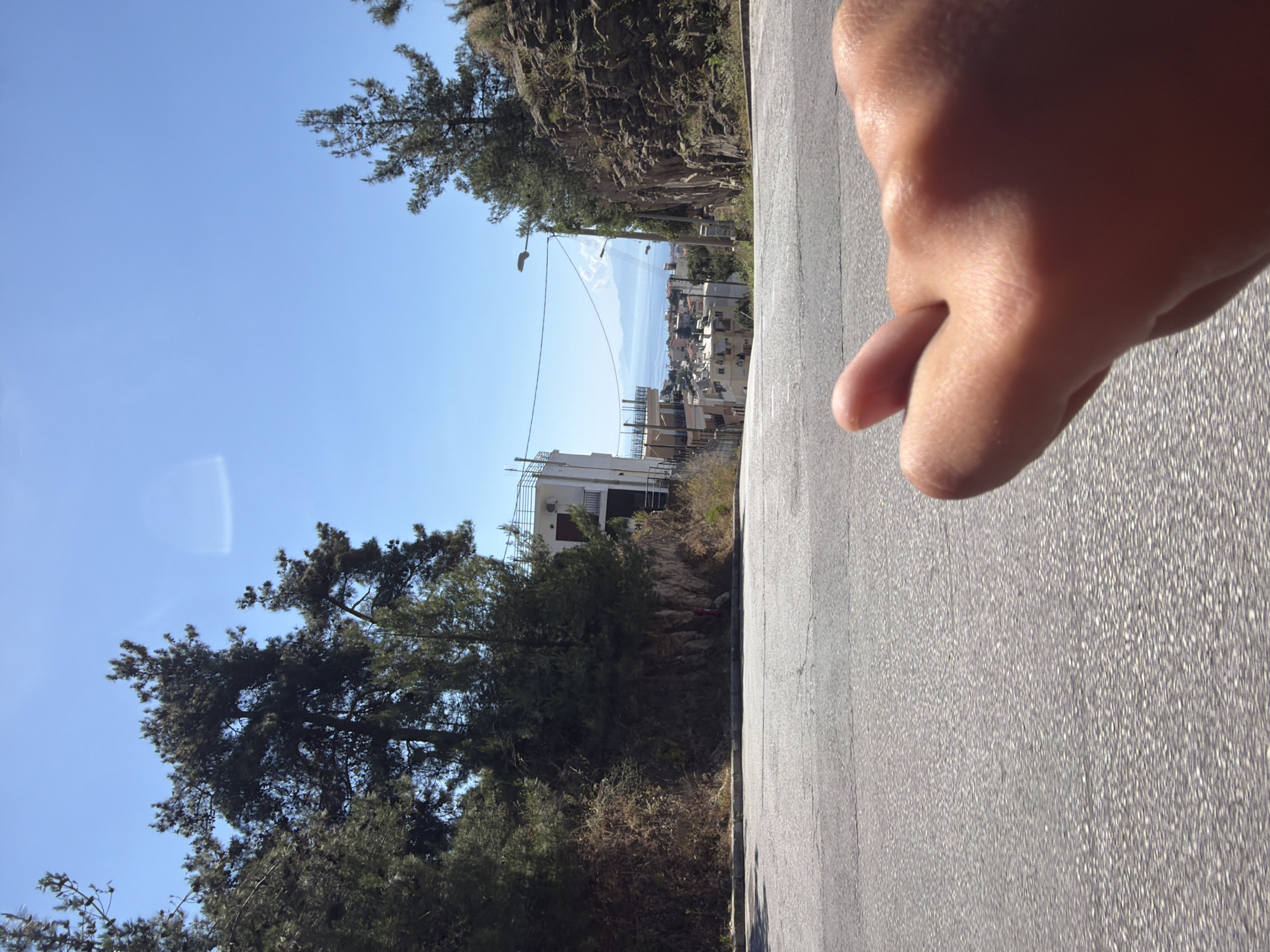 The image captures a scene from an elevated road in Kavala, Greece. The foreground is dominated by an expanse of grey asphalt, partially obscured by a human hand in the upper right corner. Beyond the road, the terrain slopes downward, with a mix of trees and vegetation visible. In the distance, a cluster of buildings indicates an urban or suburban area, possibly the city itself. A glimpse of the sea and distant mountains is visible in the background, under a clear blue sky. A few utility poles and overhead wires are seen near the buildings.