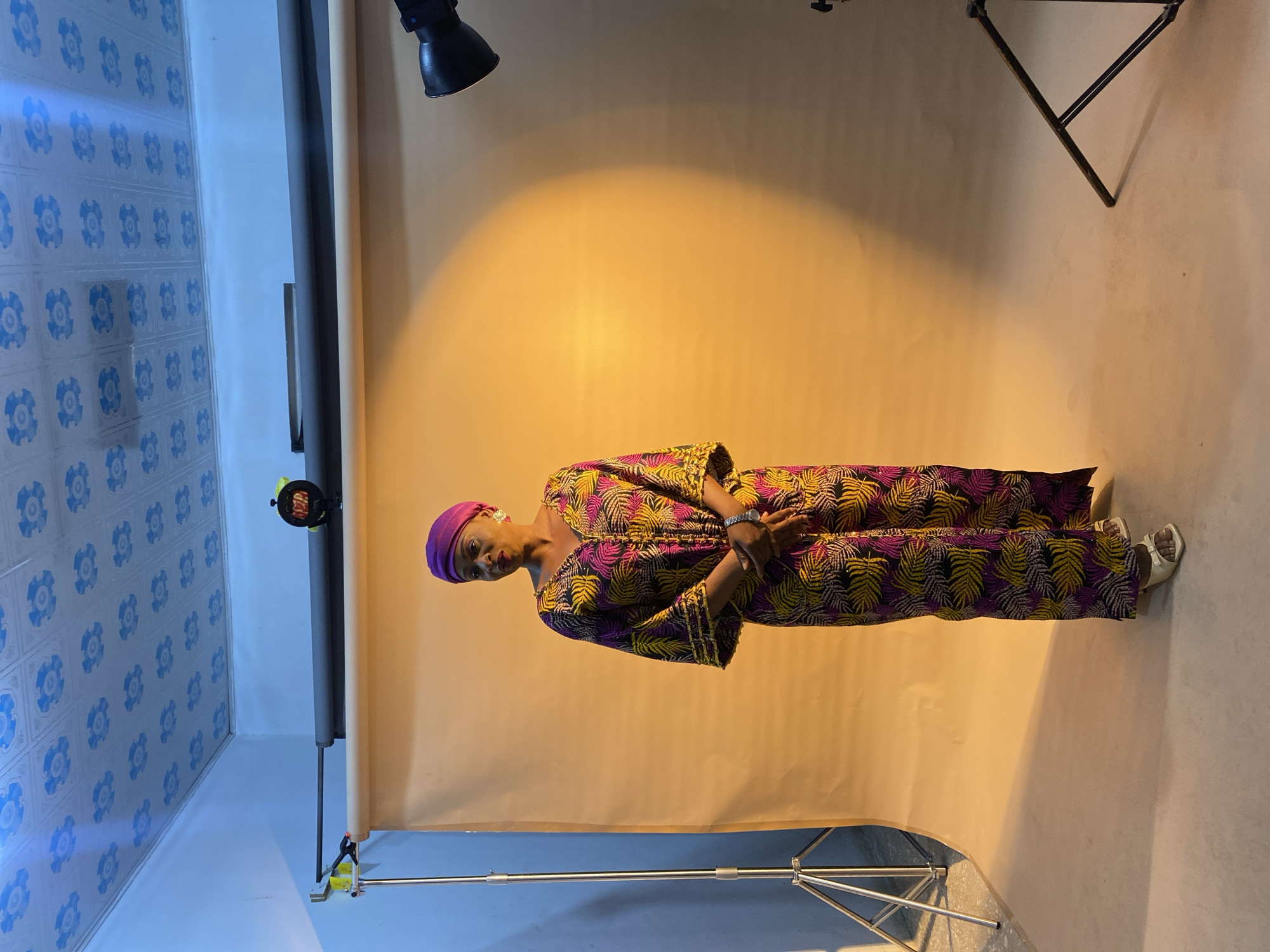 A woman is standing in a studio against a tan backdrop. She is wearing a purple head covering and a long patterned dress in purple, gold, and black. She is standing with her hands clasped in front of her. The studio has blue patterned walls on the left side of the image and lighting equipment above the woman. The city of Gura Top, Nigeria, is not discernible from the image.