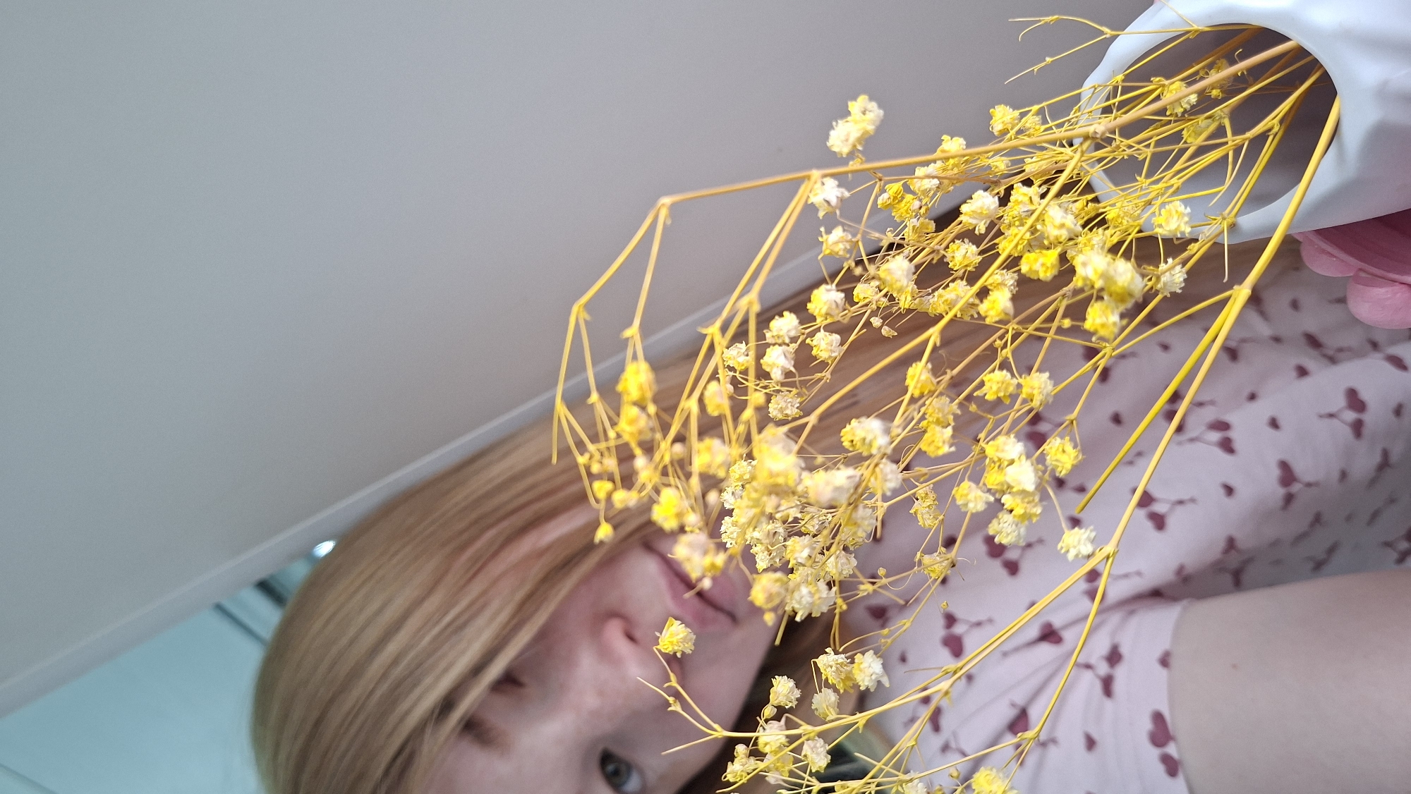 The image shows a woman with blonde hair, wearing a light pink shirt with a small floral pattern, partially visible behind a bouquet of dried yellow flowers in a white vase. The shot is taken from a low angle, looking upwards towards a white ceiling. The woman's face is partially obscured by the flowers, but her eyes and mouth are visible. The exact location within the city of Chaikovsky, Russia, is not discernible from the image.