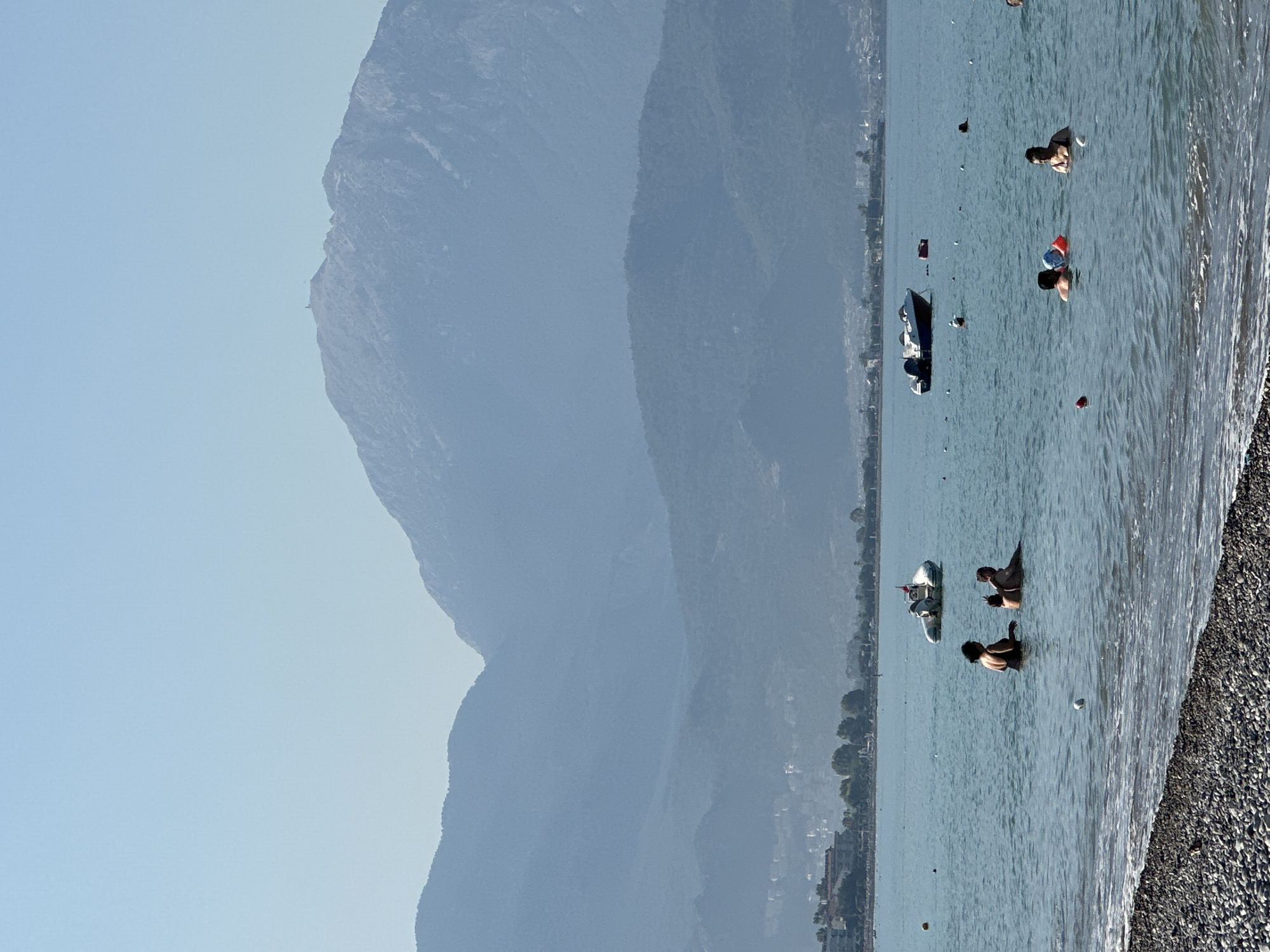 The image depicts a scenic view of the coastline in Fethiye, Turkey, with the Mediterranean Sea in the foreground and the mountains of the Lycian coast in the background. The sea is calm and a few people are seen swimming and enjoying the water. A small boat is anchored offshore, and a few buoys mark a swimming area. The beach is covered with pebbles. The distant mountains are shrouded in a light haze, and a few buildings are visible along the coastline. The sky is clear and blue, indicating a pleasant day.