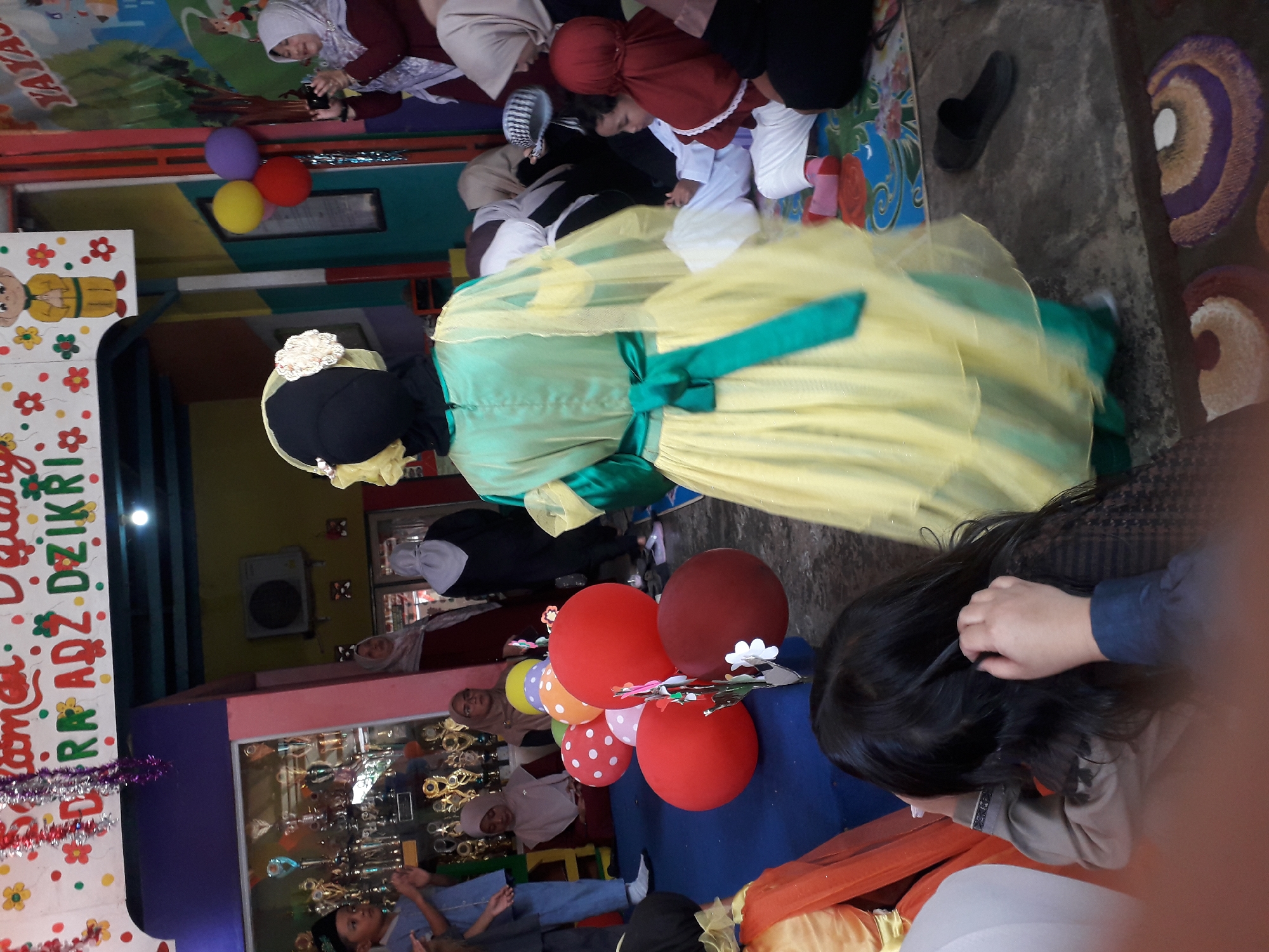 The image depicts an indoor event, possibly a school or community gathering, in Cankuang, Indonesia. In the foreground, a person in a yellow and green traditional outfit, possibly a dress or costume, stands facing away from the camera. Their back is to the viewer, and they are wearing a black head covering with a decorative white flower. To the left, a child with dark hair is partially visible, reaching up towards their head.
Behind this figure, a cluster of red, orange, yellow, and white balloons are arranged. Several children are seated on the floor in the middle ground, some wearing traditional clothing and headscarves. A young boy in a light blue outfit stands to the left, his hands clasped. In the background, a display of trophies is visible behind a glass partition. Adults are also present, some wearing hijabs, and one is seen taking a picture with a camera. The environment appears to be decorated, with colorful banners and drawings on the wall. A wall-mounted air conditioning unit is visible on the upper left. The floor is covered with mats featuring colorful patterns. A black slipper is placed on the floor to the right.