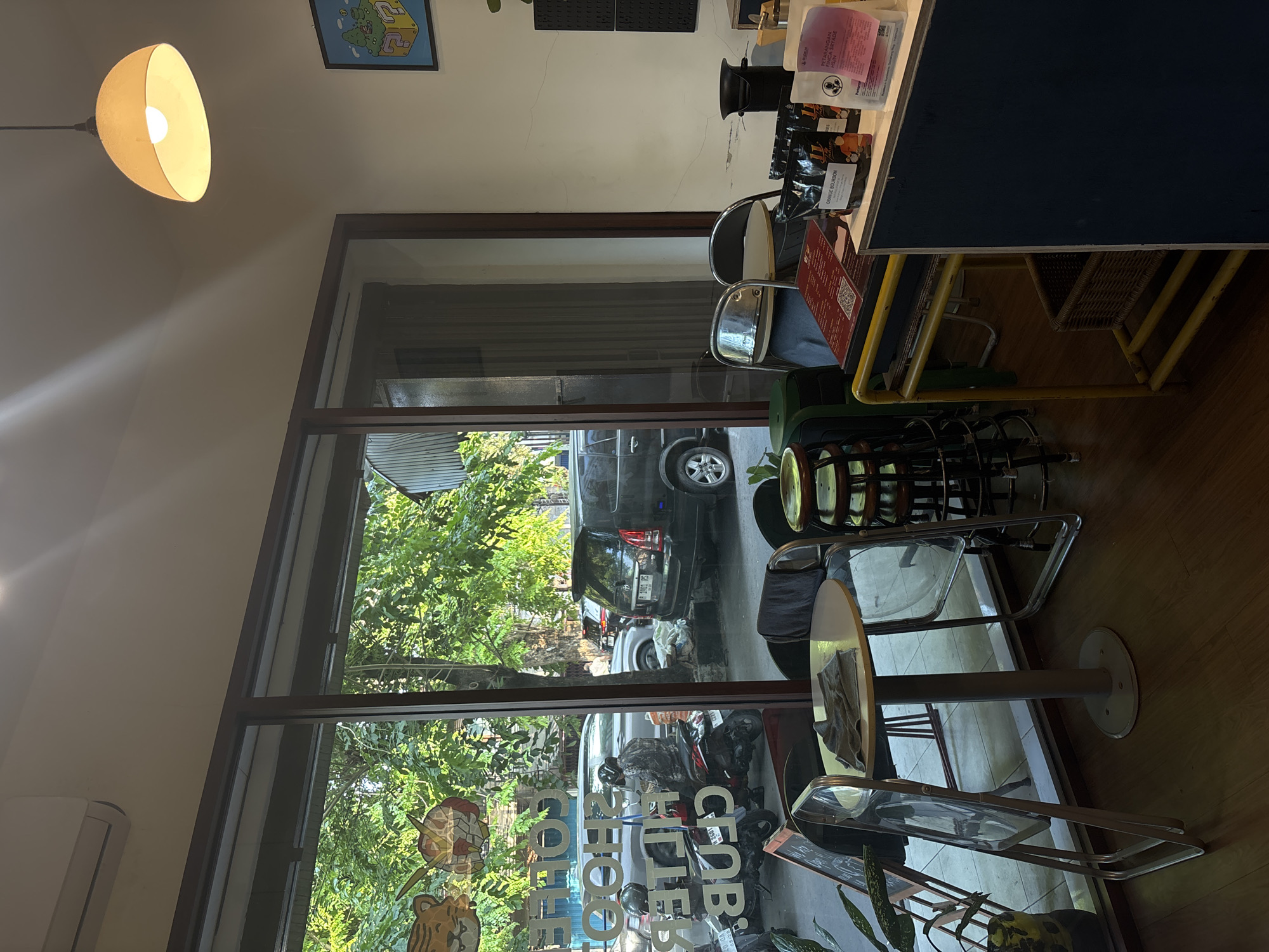 The image depicts the interior of a cafe with a large glass window overlooking a street scene in Semarang, Indonesia. Inside the cafe, there are several tables and chairs. One round table with a gray top and a central stand is visible in the foreground. Two clear plastic chairs are placed around it, one of which is partially folded. A gray cloth is draped over the table. To the right, a yellow metal cart with a woven basket underneath holds various items, including coffee bags, one labeled "ORANGE BOURBON," and a red menu or flyer. A black pegboard is mounted on the wall behind the cart. A pendant lamp with a light yellow shade hangs from the ceiling.
Through the glass window, a street scene is visible with trees, parked cars, and motorcycles. A dark-colored SUV is parked on the right side of the street, and a silver car is parked behind it. A person is seated on a red motorcycle in front of the silver car, wearing a patterned jacket and helmet. The street appears to be lined with trees, with sunlight filtering through the leaves. The overall ambiance suggests a bright, daytime setting.