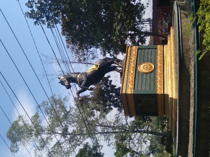 A large, dark-colored horse statue, depicted rearing on its hind legs with a gold bridle and a yellow and black decorated saddle blanket, stands prominently in an outdoor setting in Hegarmanah, Indonesia. The statue is mounted on a multi-tiered pedestal; its top and bottom sections are gold with ornate relief carvings, while the middle section is dark green with horizontal grooves. The green section features a central circular gold medallion with a radial design and an unreadable dark rectangular plaque to its left. The pedestal rests on a circular, gold-colored base with a darker concrete or stone rim, surrounded by visible ground and sparse green foliage. The background consists of a clear blue sky, partially traversed by multiple black overhead power lines. Numerous trees of varying heights and foliage density surround the monument, including tall, slender trees with light green leaves and denser, darker green trees. In the distant right background, a red and dark-colored structure with banners is partially visible.