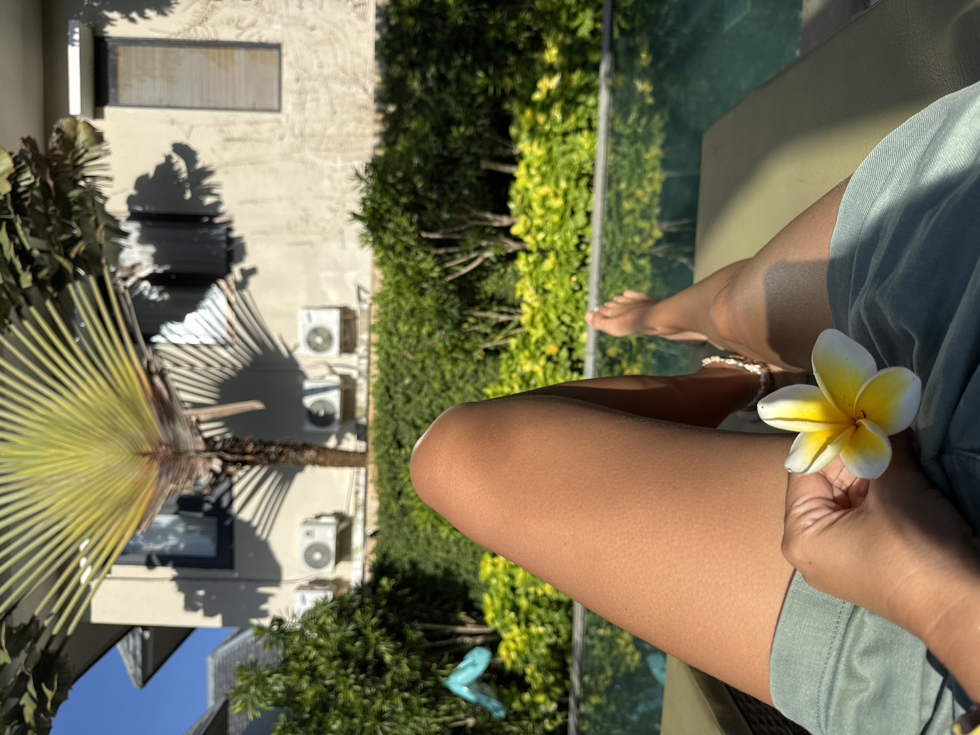 A first-person perspective shot shows a person sitting on a lounge chair, possibly by a pool. The individual's legs are visible, and they are holding a frangipani flower. In the background, there's a building with multiple air conditioning units attached to its exterior wall. Vegetation is present, including trees and bushes. The location is likely Kuta Selatan, Indonesia, given the context.