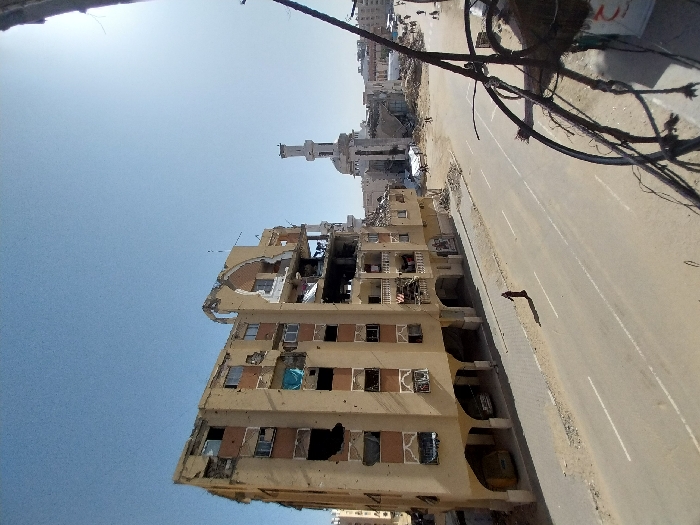 An elevated view in Khan Yunis, Palestinian Territory, reveals a severely damaged urban landscape under a clear blue sky. A prominent multi-story, light-beige building, approximately six to seven stories tall, dominates the left mid-ground, exhibiting extensive structural damage, including missing facade sections, exposed interiors, and numerous projectile impact marks on its exterior. Shattered windows and damaged balconies are visible, with some fabric hanging from one. The ground floor features arched bays, partially obscuring two vehicles. A two-lane asphalt road with white lane markings spans the lower half of the image. A lone individual, dressed in dark attire, walks on the road, casting a shadow. In the background, a white mosque with a tall minaret stands amidst widespread rubble and debris from collapsed buildings. Additional damaged structures and small human figures are discernible in the far distance. Tangled wires occupy the immediate right foreground, suggesting the vantage point is from a damaged structure.