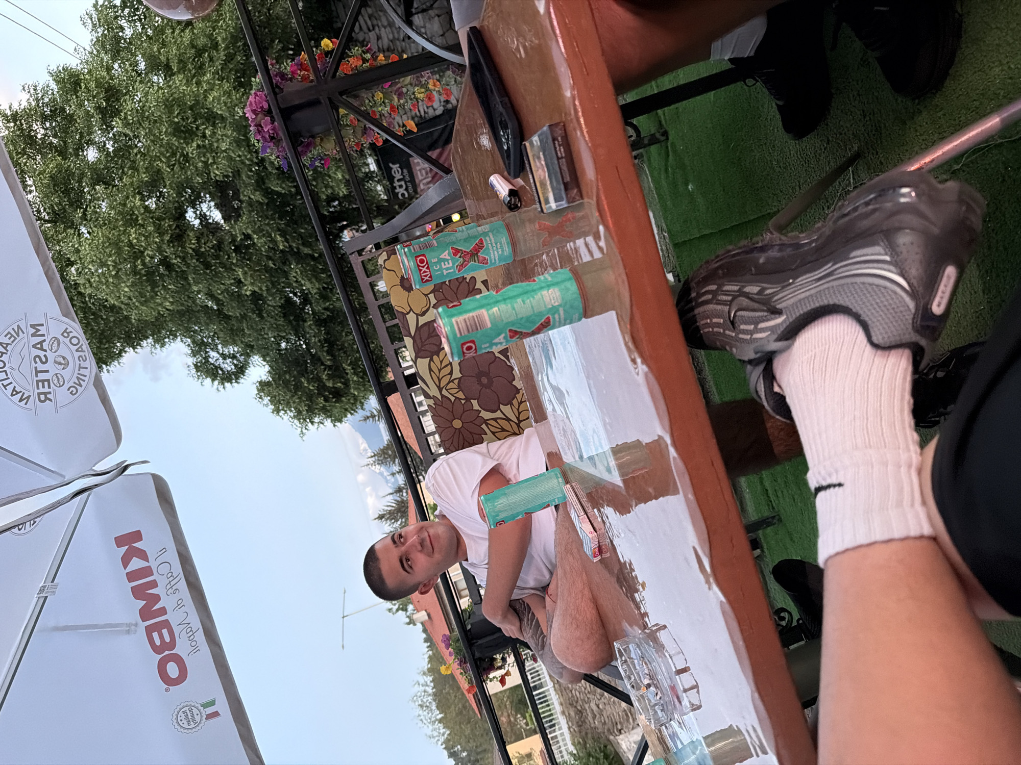 The scene captures a man at an outdoor cafe, positioned near a table with cans of Xixo Ice Tea and smoking accessories. He is wearing a white t-shirt, with part of an umbrella bearing the word "KIMBO" visible above him. A flower arrangement hangs from the cafe's awning, adding a touch of nature to the urban setting. Another person, partially visible with a black shoe, white socks, and a dark-colored upper garment, suggests a shared setting. The scene is well-lit, with outdoor elements like a tree and a glimpse of a fence in the background.