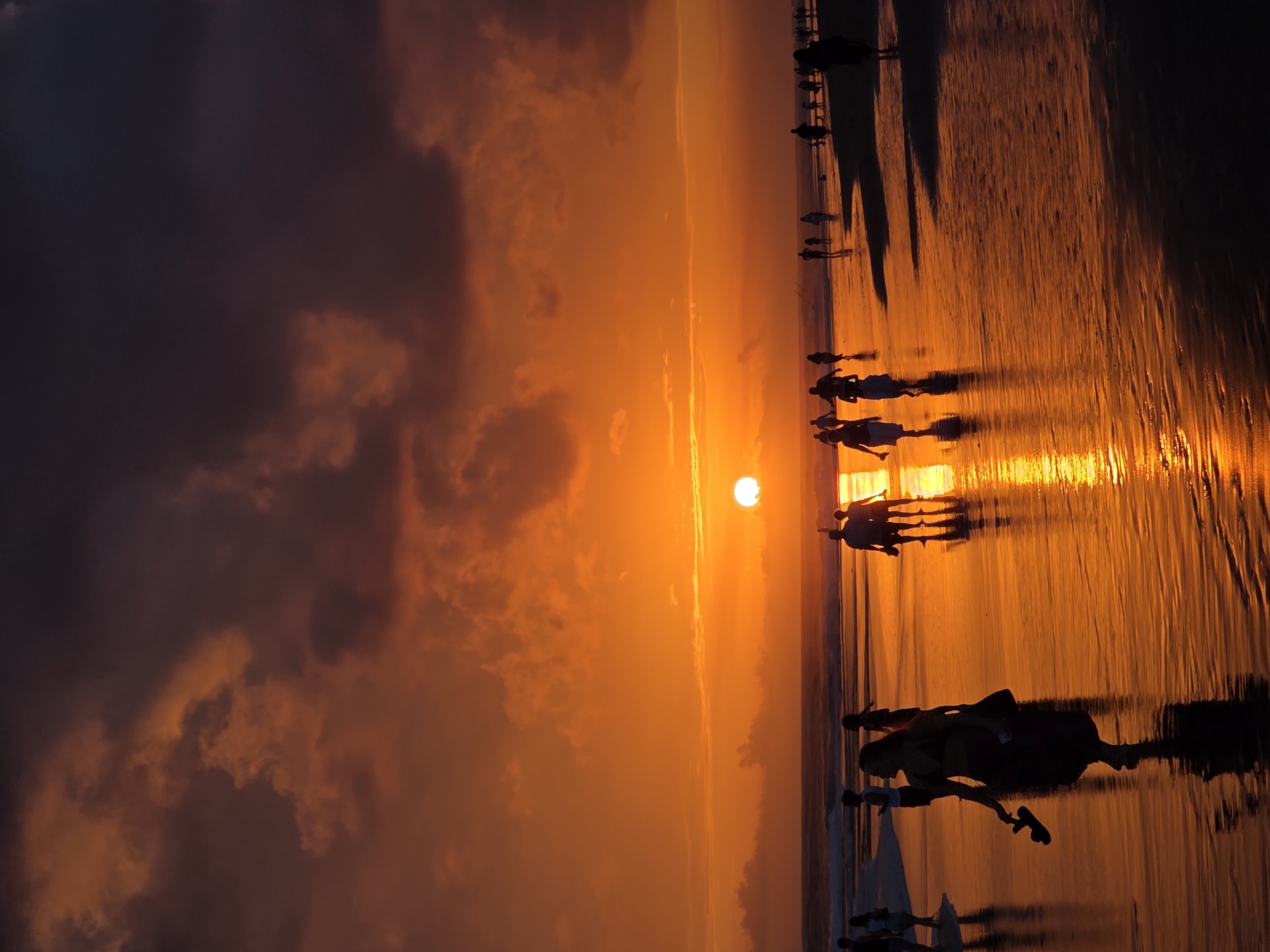 The image depicts a vibrant sunset scene on a beach in North Kuta, Indonesia. Silhouetted figures of people are scattered across the wet sand, reflecting the warm hues of the sky. The sun, a bright orb of light, is positioned near the horizon, casting a golden glow across the ocean and the beach. Dark clouds dominate the upper portion of the sky, creating a dramatic contrast with the fiery orange and yellow tones below. Waves gently lap at the shore, adding to the serene atmosphere. A woman in the foreground is walking along the beach, holding her shoes, with her reflection visible on the wet sand. Several other individuals and groups are visible in the distance, enjoying the evening spectacle. The overall impression is one of a tranquil and picturesque beach setting at dusk.