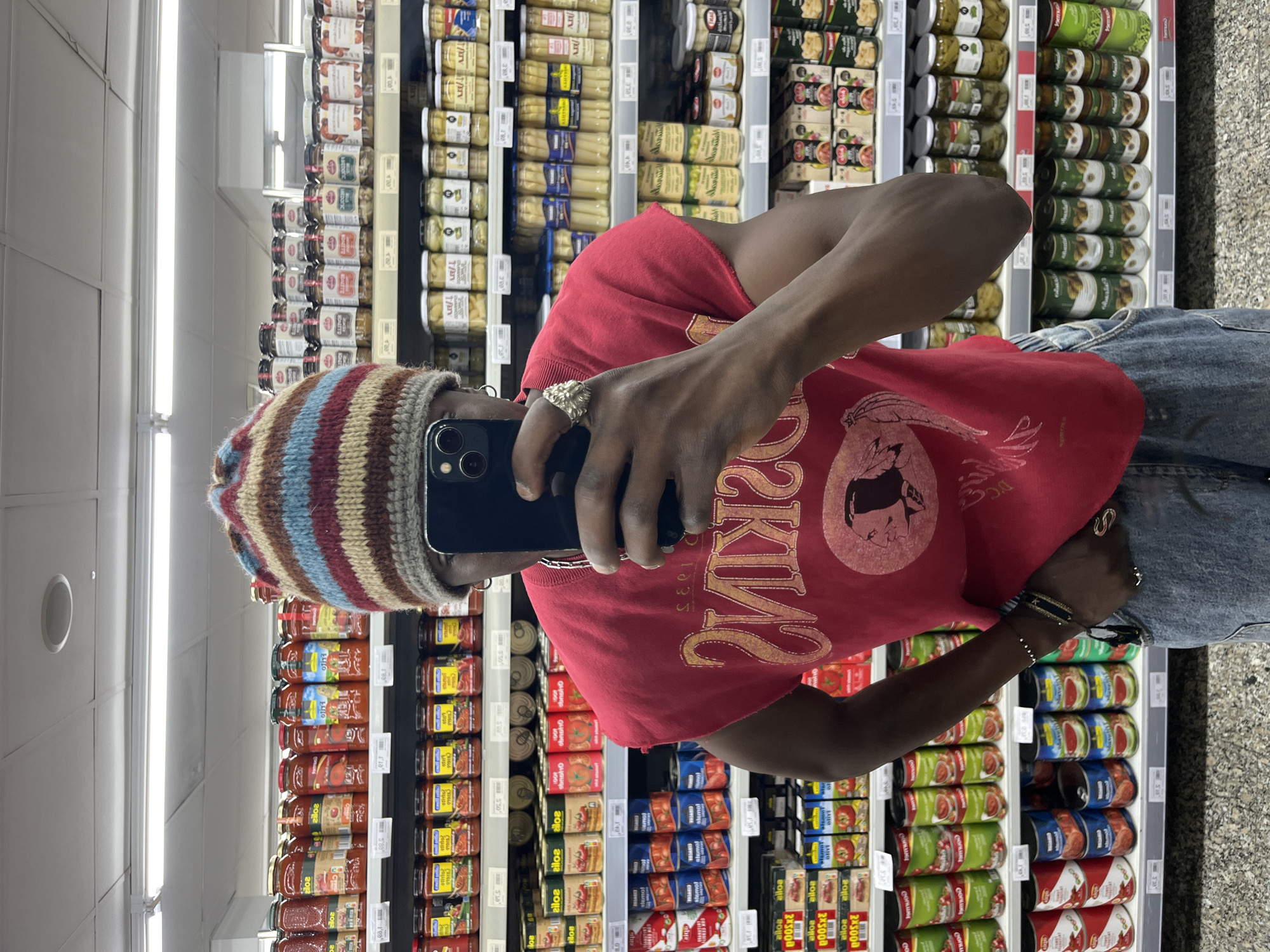 The image, which appears to be rotated 90 degrees counter-clockwise, depicts a person taking a mirror selfie inside a supermarket aisle. The individual, seen from the chest up, holds a dark-colored smartphone that obscures their face. They are dressed in a red t-shirt featuring a faded graphic with visible text including "2KIN", "D'C", and "1935". The outfit is completed with blue denim jeans and a multi-colored striped beanie in shades of brown, blue, red, and beige. Accessories include a prominent silver ring on the right index finger, additional rings, and multiple bracelets on the left wrist.
The background shows brightly lit supermarket shelves filled with various canned and jarred goods, characteristic of a grocery store environment, likely located in Barcelona, Spain, as per the provided context. The shelves display products such as preserved asparagus, olives, mushrooms, and tomato sauces. Some discernible text on product labels includes "alloa", "Contu", "Jomuo", "zile", "obnoho", "silon", "stemol", and "AR/L". Price tags or product codes like "5'50", "198", and "10" are also visible among the merchandise. The scene is illuminated by artificial fluorescent lighting, typical of an indoor retail space.