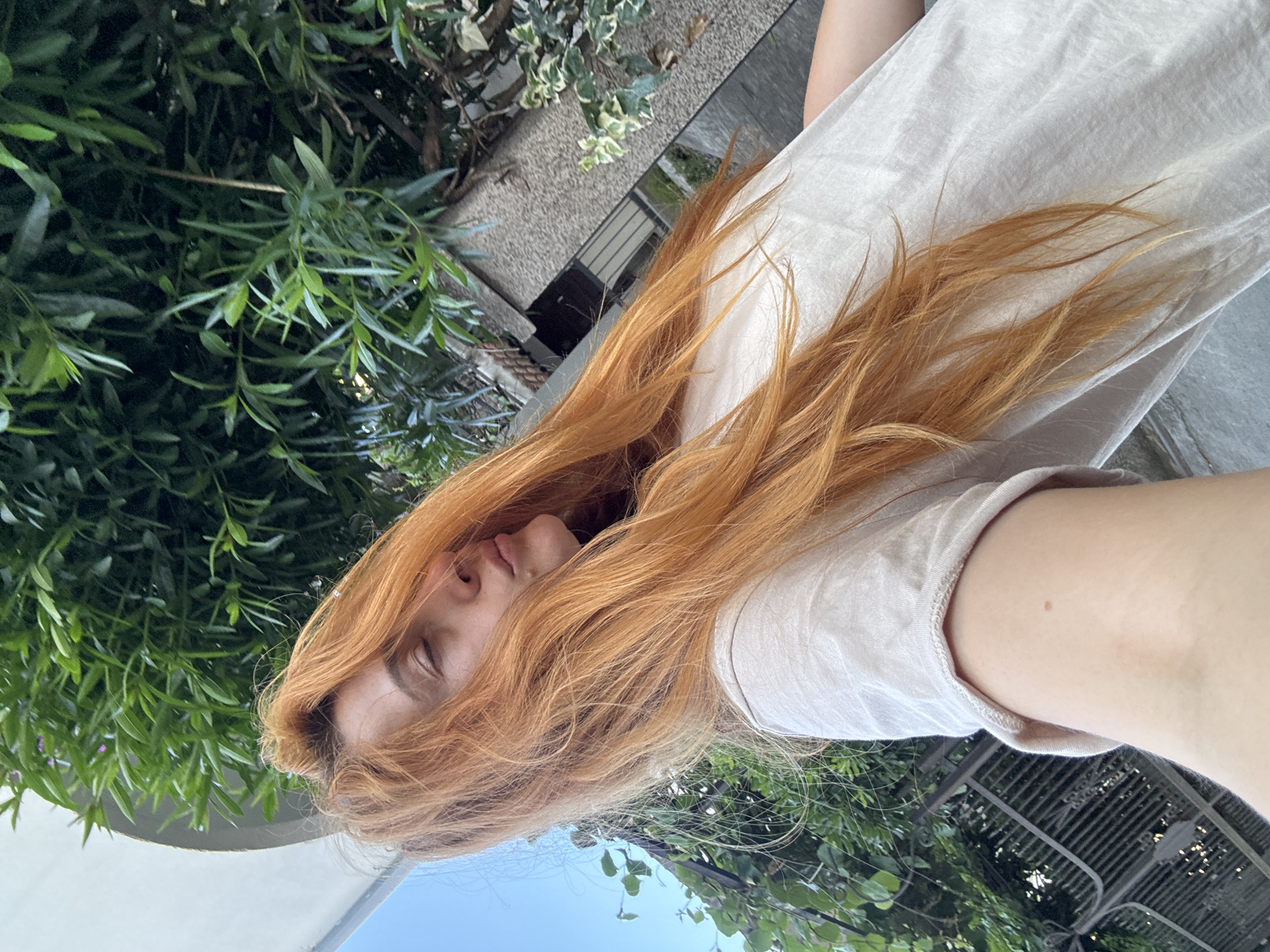 A person with long, wavy, reddish-orange hair is lying down outdoors, with their head and upper body dominating the frame. Their eyes are closed and their mouth is slightly open, suggesting a relaxed or perhaps sleepy state. They are wearing a light-colored, loose-fitting t-shirt. The background consists of lush green foliage, a concrete structure that appears to be part of a building, and a clear blue sky. The image is captured from a high angle, looking down on the person. The specific location in the city of Budva, Montenegro cannot be determined from the visual information provided.