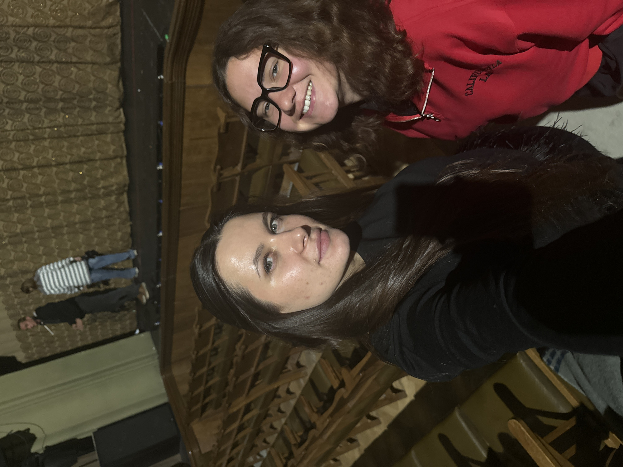 Two women take a selfie in the Bolshoi Theater in Moscow, Russia. One woman wears a red sweatshirt and glasses. The other wears a black top. The theater's architecture and seating are visible in the background. In the reflection of the building's windows, two people can be seen. One wears a striped sweater.