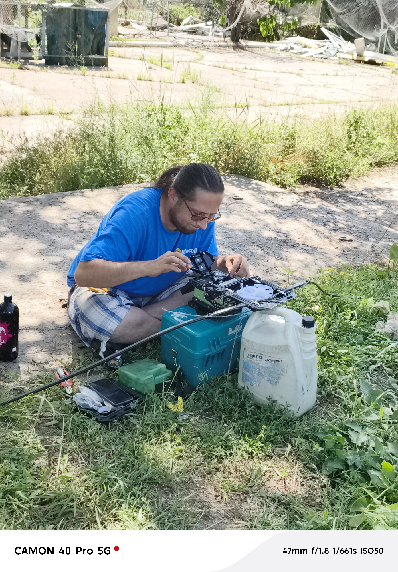 A man wearing glasses and a blue t-shirt with the logo "lifecell" is sitting on the ground in an outdoor, grassy area. He is focused on working with a piece of equipment that appears to be for fiber optic splicing, with wires and tools laid out around him. To his left, a dark bottle with a pink label is visible. In front of him, a teal-colored toolbox and a large white plastic jug are on the ground. The background shows a dry, cracked ground surface with sparse vegetation and some debris, suggesting an industrial or undeveloped area. The scene is illuminated by natural sunlight, casting shadows on the ground.