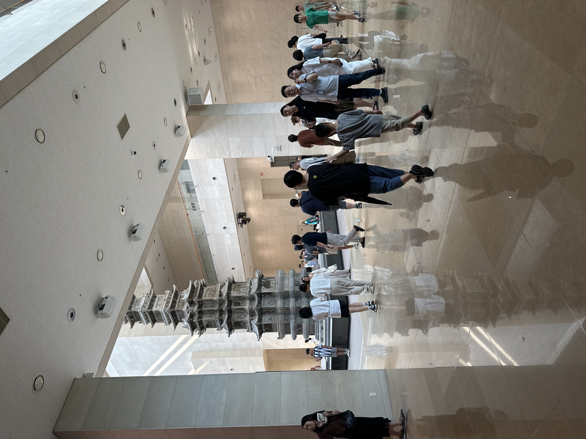 The image depicts the interior of a museum lobby, likely in Seoul, South Korea, given the presence of a large stone pagoda. The space is expansive with high ceilings and polished marble floors that reflect the overhead lighting and the surrounding architecture. Natural light streams in from a large window or opening visible in the background, illuminating the area.
Dominating the center of the frame is a tall, intricately carved stone pagoda, appearing to be an artifact on display. Several individuals are visible throughout the lobby, appearing to be museum visitors. They are casually dressed and engaged in various activities such as walking, observing the pagoda, and conversing.
In the foreground, a woman in a dark dress is standing to the left, holding a small bag and looking to her left. Towards the center, a group of people are walking away from the camera, including a young boy in a striped shirt and shorts. Further back, more people are milling about, some in groups, others individually.
The environment suggests a well-maintained public space, likely an art museum or cultural institution, given the architectural style and the prominent display of the pagoda. The overall atmosphere is one of quiet observation and movement within a large, open hall.