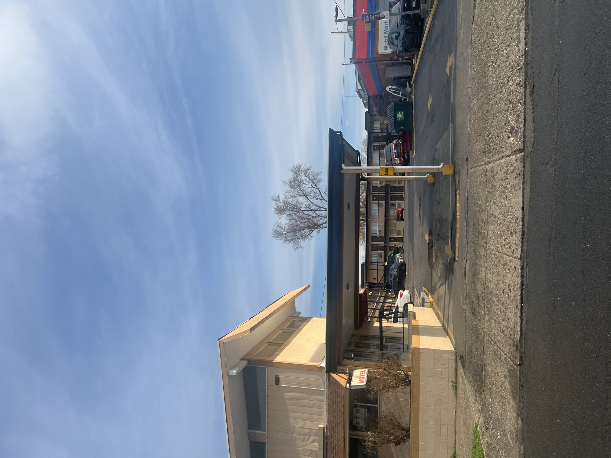 The image shows a motel in the City of Newburgh, United States of America. The motel is a two-story building with beige walls and a brown roof. There is a sign that says "MOTEL" on the front of the building. There is a Mobil Mart gas station in the background. The sky is blue and there are some clouds.
