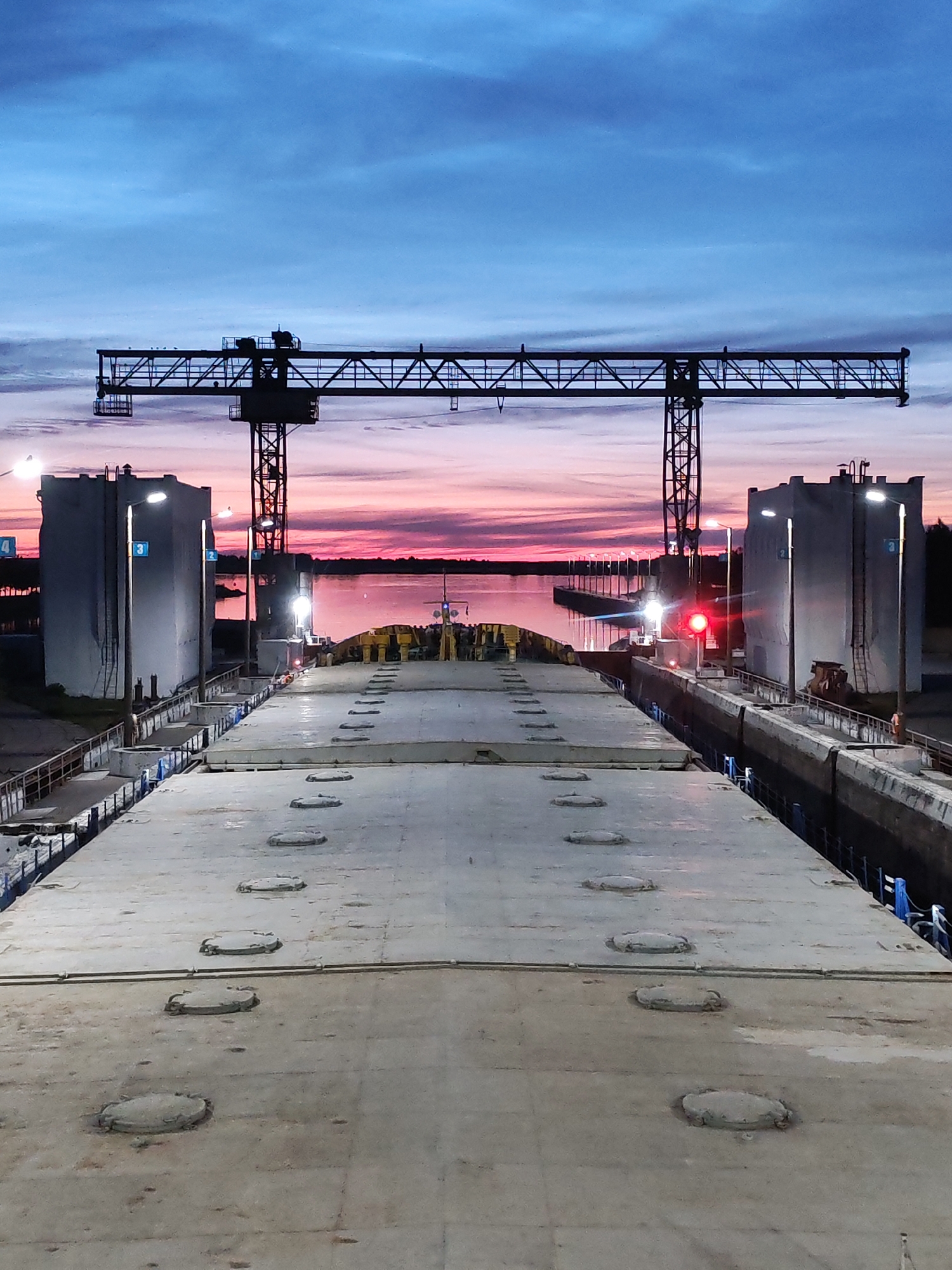 The image captures a twilight scene at a lock on the Sheksna River in Sheksna, Russia. A large, flat-topped barge is docked within the lock. The sky displays a gradient of deep blue overhead, transitioning to vibrant pink and orange hues near the horizon, indicative of sunset or sunrise. Towering over the lock are large, industrial gantry cranes, silhouetted against the colorful sky. On either side of the lock are imposing, rectangular structures, possibly control buildings or lock gates, illuminated by overhead lights. Several smaller lights are visible along the water's edge. The water surface reflects the dramatic sky. The barge itself is concrete-colored and shows circular, recessed hatches distributed across its deck. The overall impression is one of industrial activity at the confluence of man-made engineering and natural light.