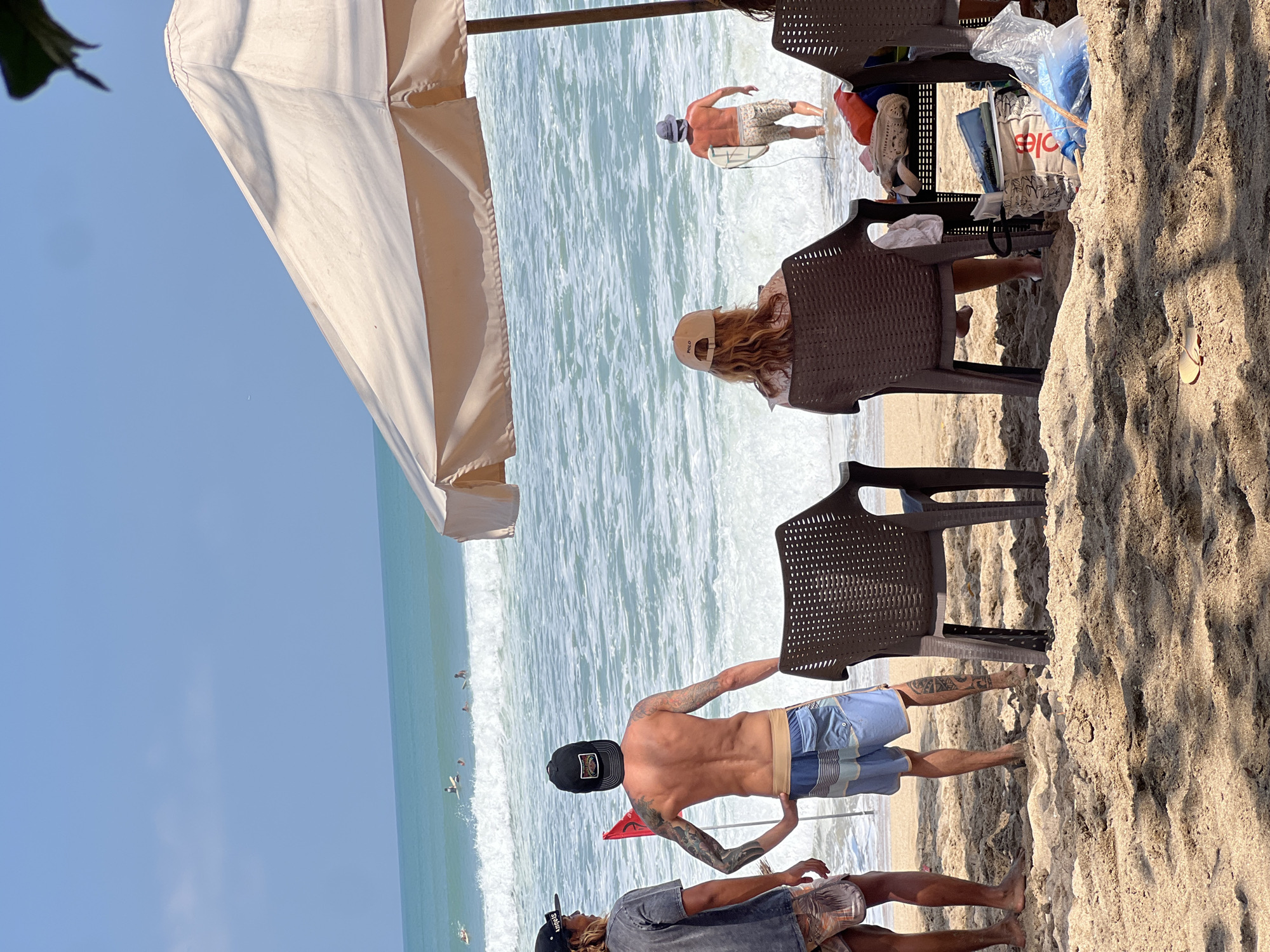 The image depicts a sunny day at Kuta Beach, Bali, Indonesia. In the foreground, three individuals are seated on plastic chairs under a large, light-colored beach umbrella, facing the ocean. Their backs are to the camera. The person in the center is a woman wearing a light-colored shirt and a tan baseball cap. To her left is another person whose face is obscured. To her right, another person's arm is visible.
Further back, two men stand on the wet sand near the water's edge, also facing the ocean. The man on the left is wearing a dark t-shirt and patterned shorts, with a black baseball cap. The man on the right is shirtless, wearing blue and white striped board shorts and a black baseball cap. He has tattoos on his arms and back. Between them is a red flag planted in the sand.
In the water, several surfers are visible on their boards, riding waves. Another man, wearing a hat and patterned shorts, walks into the ocean carrying a surfboard. The turquoise water is choppy with white foam from the waves. The sky is clear and blue. The sand in the foreground is light brown and textured. Items like bags and a red container are visible on the ground near the seated individuals.