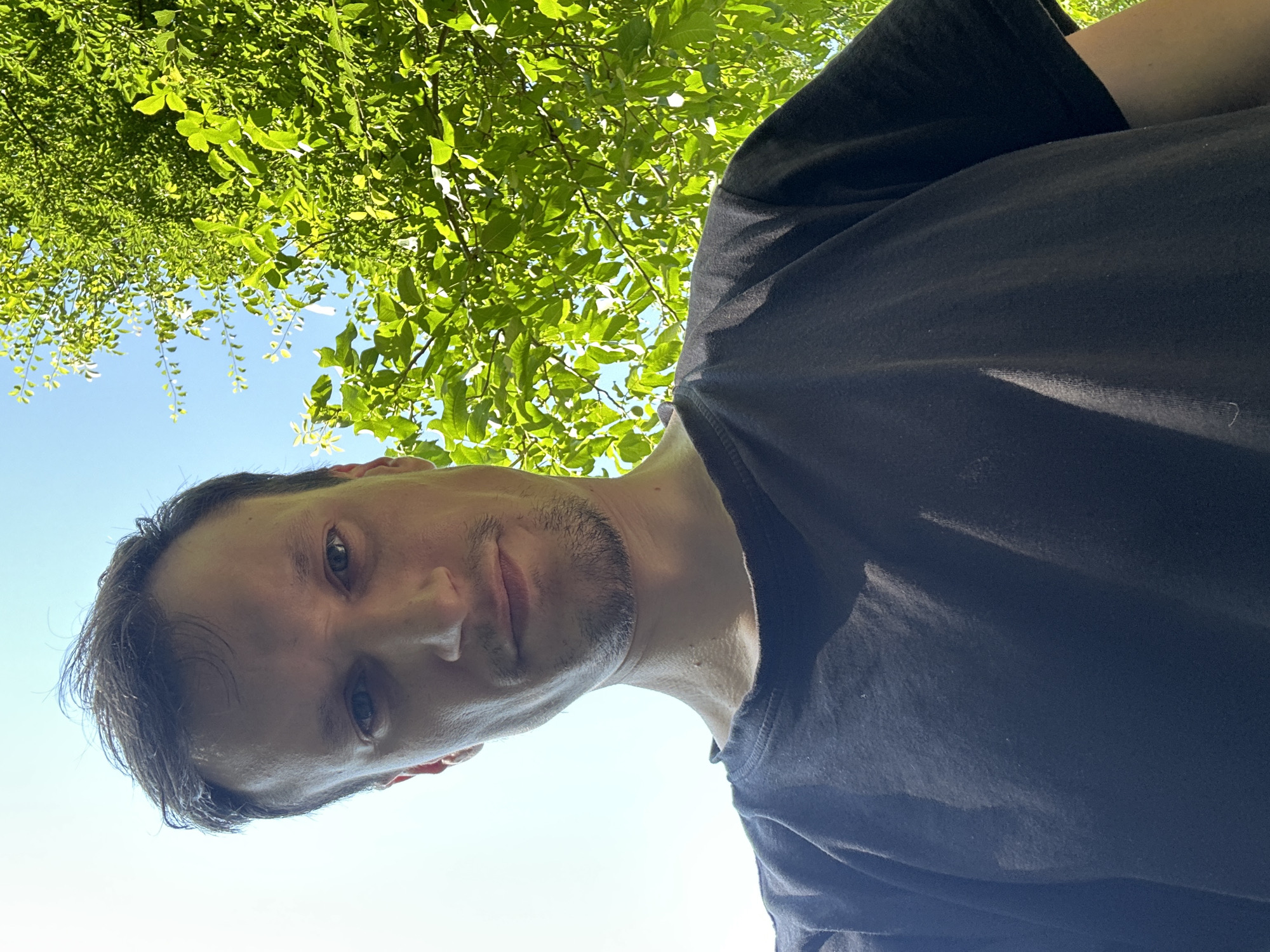 A low-angle, close-up selfie of a man is taken outdoors against a bright blue sky and vibrant green foliage. The man, who appears to be in his late 20s or early 30s, has light skin, short brown hair, and a light beard and mustache. He is wearing a dark t-shirt. The sunlight casts shadows across his face and shirt. The background consists of lush green leaves and branches of a tree, with patches of blue sky visible through the canopy. The image does not provide any information about the location within the city of Makiv, Ukraine.