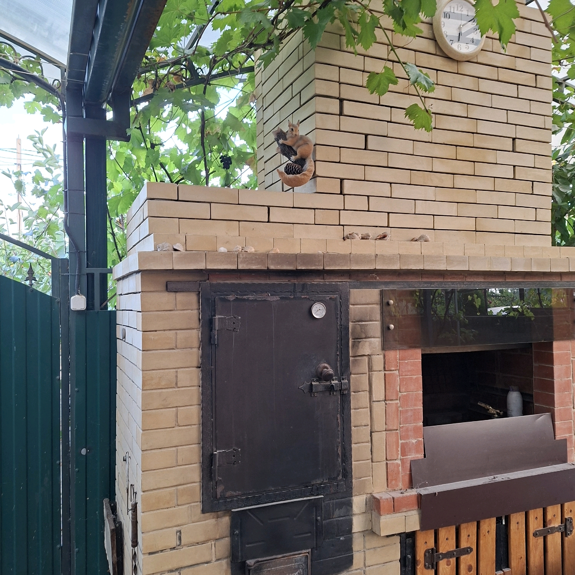 This is a medium shot of an outdoor brick oven, likely a barbecue or pizza oven, located in Anapskaya, Russia. The oven is constructed from light beige bricks and has a dark metal door with a thermometer and ornate latches. Above the door, a small decorative squirrel figurine is perched on a branch. To the right of the door is a glass panel revealing the oven's interior. A clock with Roman numerals is mounted on the brickwork above the glass panel. Green vines with leaves and some dark grapes hang over the oven from an overhead structure, suggesting a vineyard or garden setting. To the left of the oven is a dark green corrugated metal fence. The overall scene depicts a functional outdoor cooking area in what appears to be a residential or dacha setting.