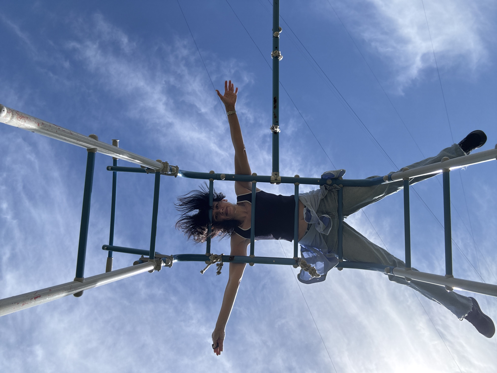A low-angle, wide shot shows a person suspended from a metal structure against a bright blue sky with wispy clouds. The person, who appears to be a woman with dark, wind-blown hair, is upside down, with her arms and legs extended. She is wearing a dark tank top, light-colored jeans, and dark shoes. One arm is raised, with her hand open as if waving, while the other arm hangs down, with her hand reaching towards the bottom of the frame. Her legs are spread apart, with one foot resting on a horizontal bar and the other dangling. The metal structure is made of interconnected bars and poles, painted green and silver, and appears to be part of a playground or outdoor fitness equipment. The sun is bright, casting shadows and highlighting the details of the structure and the person's clothing. The overall mood is one of freedom and exhilaration. The time of day appears to be late morning or early afternoon, given the bright sunlight and the clear sky. There are no visible text or location cues in the image, other than the general context of an outdoor recreational area.