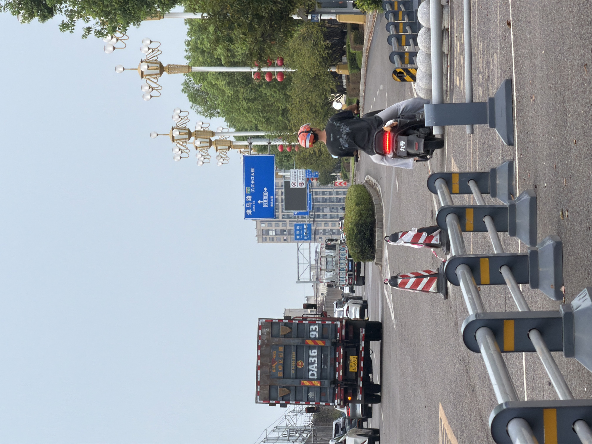 A view of a road in Jiangjin, China, shows traffic and urban elements. A truck with the license plate "DA36 93" is in the foreground, followed by a person on a scooter wearing a helmet. Traffic cones and barriers line the road, indicating ongoing construction or maintenance. Streetlights and trees are visible along the roadside, and a blue road sign indicates directions to "Jingma Rd" and "Nanshan Avenue." The sky is overcast.