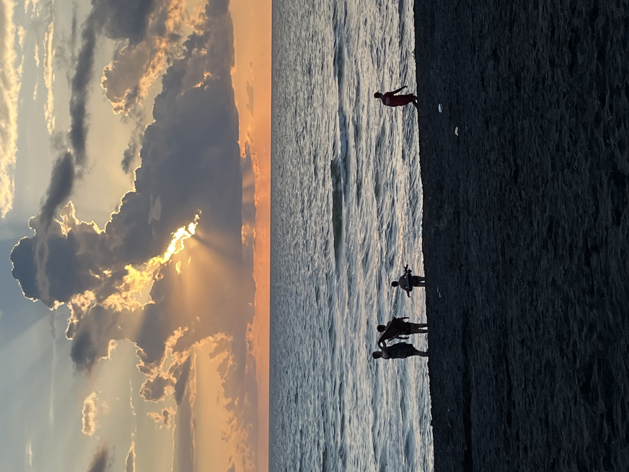 In Ureki, Georgia, the sun sets over a dark sand beach and the Black Sea. The sky is a dramatic canvas of orange and yellow hues, with dramatic clouds partially obscuring the sun, creating visible rays of light. The sea is a deep blue with gentle waves. Silhouetted figures are present on the beach. In the foreground on the left, three people stand near the water's edge, one holding what appears to be a towel or piece of cloth. To the right of this group, another person is walking along the shoreline. In the far distance on the right, a single figure in red is walking away from the camera. The beach is covered in dark, fine-grained sand.