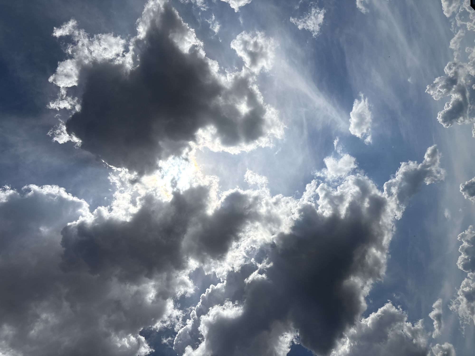The image shows a dramatic sky over Sofia, Bulgaria, with a mix of dark, heavy cumulus clouds and brighter, scattered clouds against a blue sky. The sun is partially obscured by the dense clouds, creating streaks of light and shadow that illuminate the cloud formations. The clouds are the primary subject, filling the entire frame. The lighting suggests it is daytime, possibly late afternoon or early evening, given the intensity of the sunlight breaking through. There are no visible people, buildings, or other man-made objects in the scene. The overall impression is one of dynamic weather patterns.