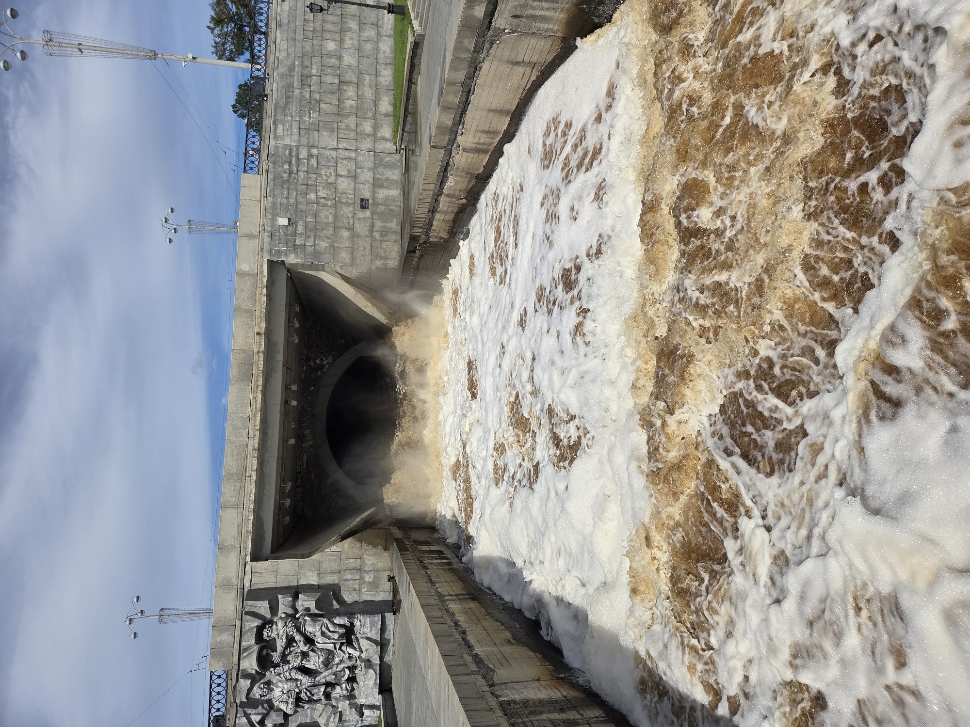 This image captures a dramatic scene of water gushing from a large concrete tunnel into a wide channel, creating turbulent, foamy rapids. The tunnel appears to be a drainage or spillway, possibly part of a dam or flood control system. The water is a murky brown, suggesting it is carrying sediment.
On the left side of the image, a large, ornate bas-relief sculpture is mounted on a concrete wall. The sculpture depicts several figures in a dynamic, historical style. Above the tunnel, a concrete bridge or elevated walkway spans the channel, supported by robust concrete structures. The surroundings suggest an urban environment, with paved walkways, lampposts, and railings visible. The sky is partly cloudy, with patches of blue sky and white clouds.
This scene is located on the Iset River embankment in Yekaterinburg, Russia, specifically near the dam of the Verkh-Isetsk Pond. The sculpture on the left is part of the monument dedicated to the founders of the city. The large tunnel is likely an overflow spillway from the dam, and the gushing water indicates a high water level or controlled release, common during spring floods or heavy rainfall in Yekaterinburg.