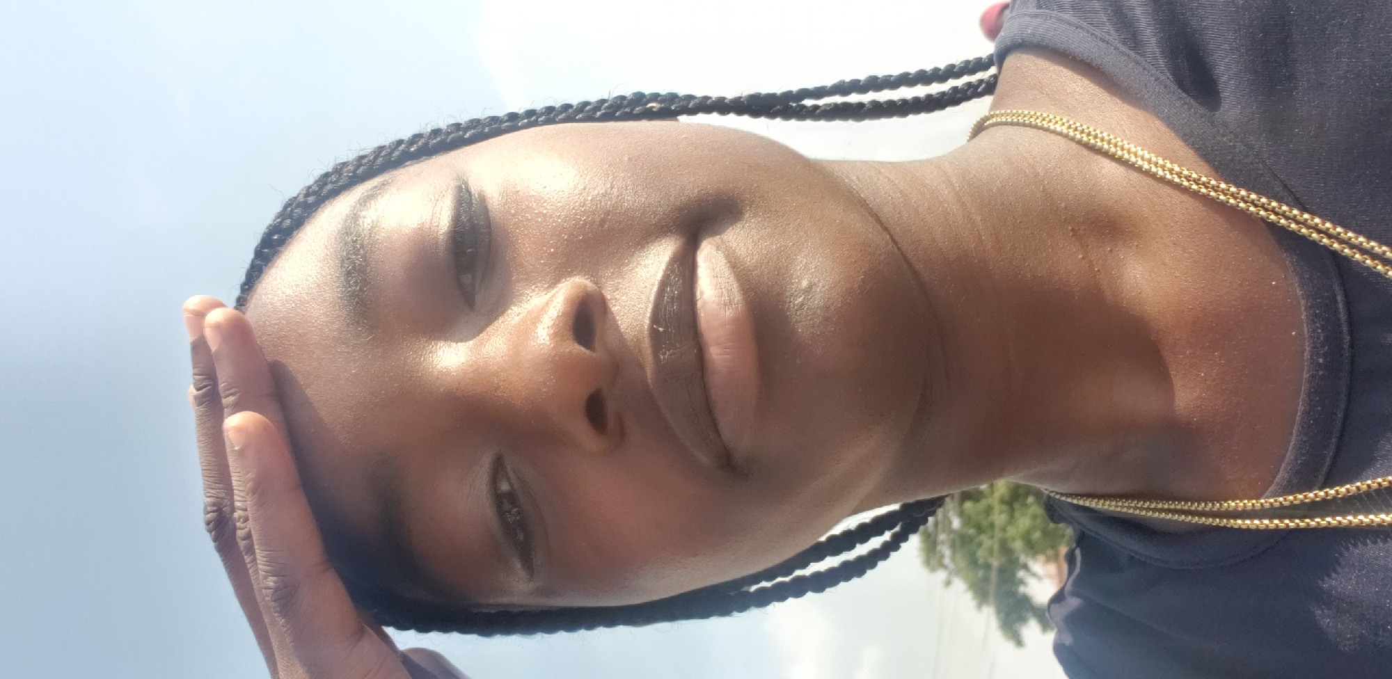 This vertically oriented image, rotated 90 degrees clockwise, features a person with dark skin and black braided hair. The subject's face is central, slightly angled upwards, with their left hand raised, fingers resting on their forehead, seemingly shielding their eyes from direct sunlight. Their expression is relaxed, with partially closed eyes and a subtle, slight smile.
The bright daytime sun creates strong highlights on the person's skin, particularly on the forehead, nose, cheeks, and lips, giving a shiny, possibly sweaty appearance. They are wearing a dark, likely navy or black, t-shirt, and a thin, double-stranded gold chain is visible around their neck and on their shoulder.
The background consists primarily of a bright, clear blue sky. In the bottom right corner of the image (which would be the lower scene when viewed upright), blurred green foliage from trees is visible, indicating an outdoor setting. The strong natural light confirms it is daytime. The overall mood conveyed is calm and contemplative. The location is Barnawa, Nigeria. No visible text is present.