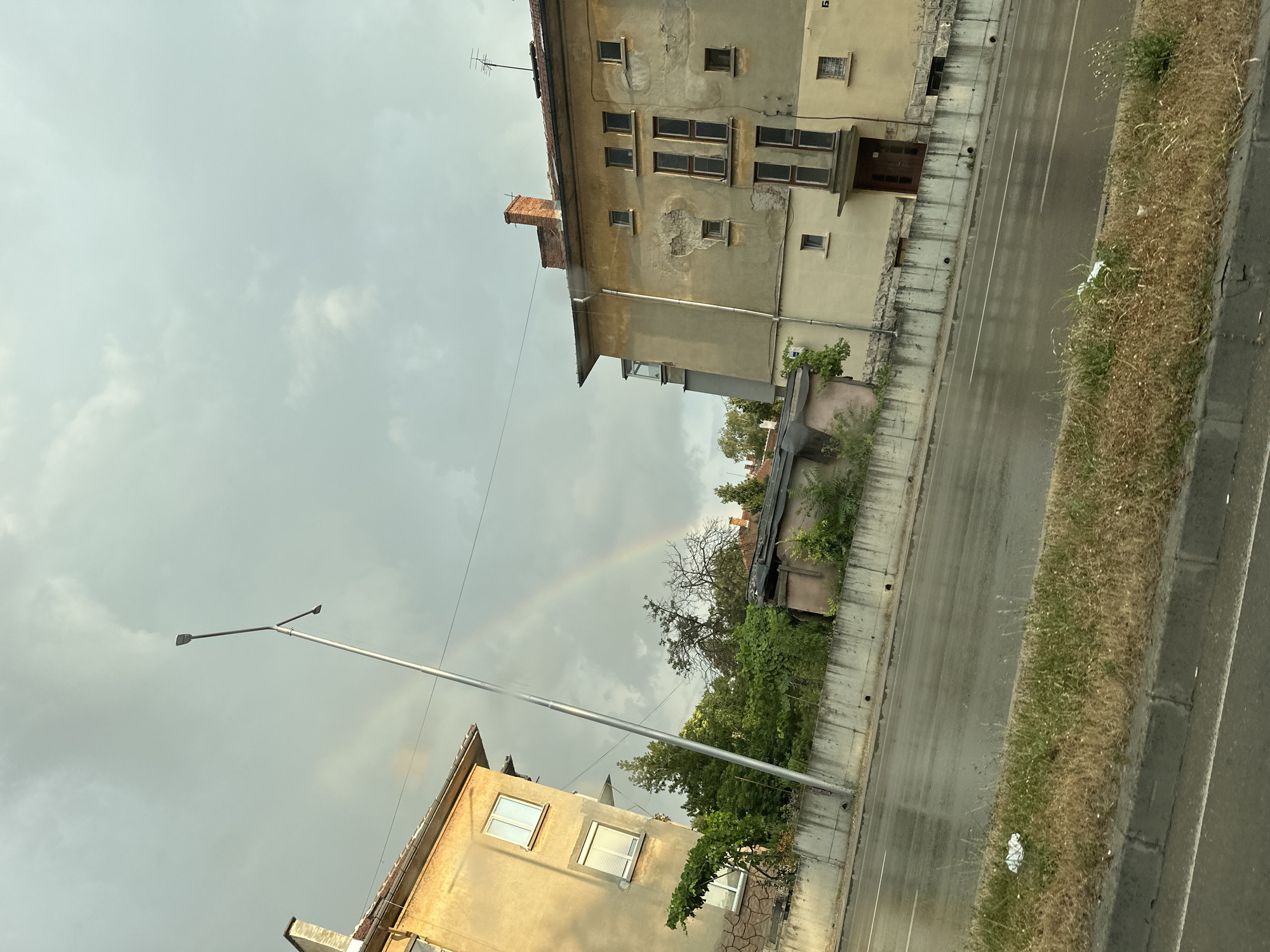 This is a vertical, eye-level, outdoor shot of an urban scene in Ruse, Bulgaria. The image captures a street with two older apartment buildings lining one side, and a grassy verge alongside a wet asphalt road on the other. The sky is overcast and grey, with a faint rainbow visible in the distance, suggesting recent or ongoing rain.
The buildings appear to be old, with weathered facades, multiple windows, and some exposed brickwork on one of them. One building has a satellite dish on its roof, and a lamp post stands tall in front of it. Green trees and bushes are interspersed between the buildings and the road. The road itself is damp and reflects the overcast sky, indicating it might have recently rained. A small amount of trash is visible on the grassy verge next to the road.
The time of day is difficult to determine due to the overcast conditions, but the presence of a rainbow suggests it is daytime, possibly late afternoon or early evening, after a rain shower. The overall mood is somewhat somber due to the grey sky, but the faint rainbow offers a hint of hope or beauty amidst the urban environment. There is no visible text in the image.